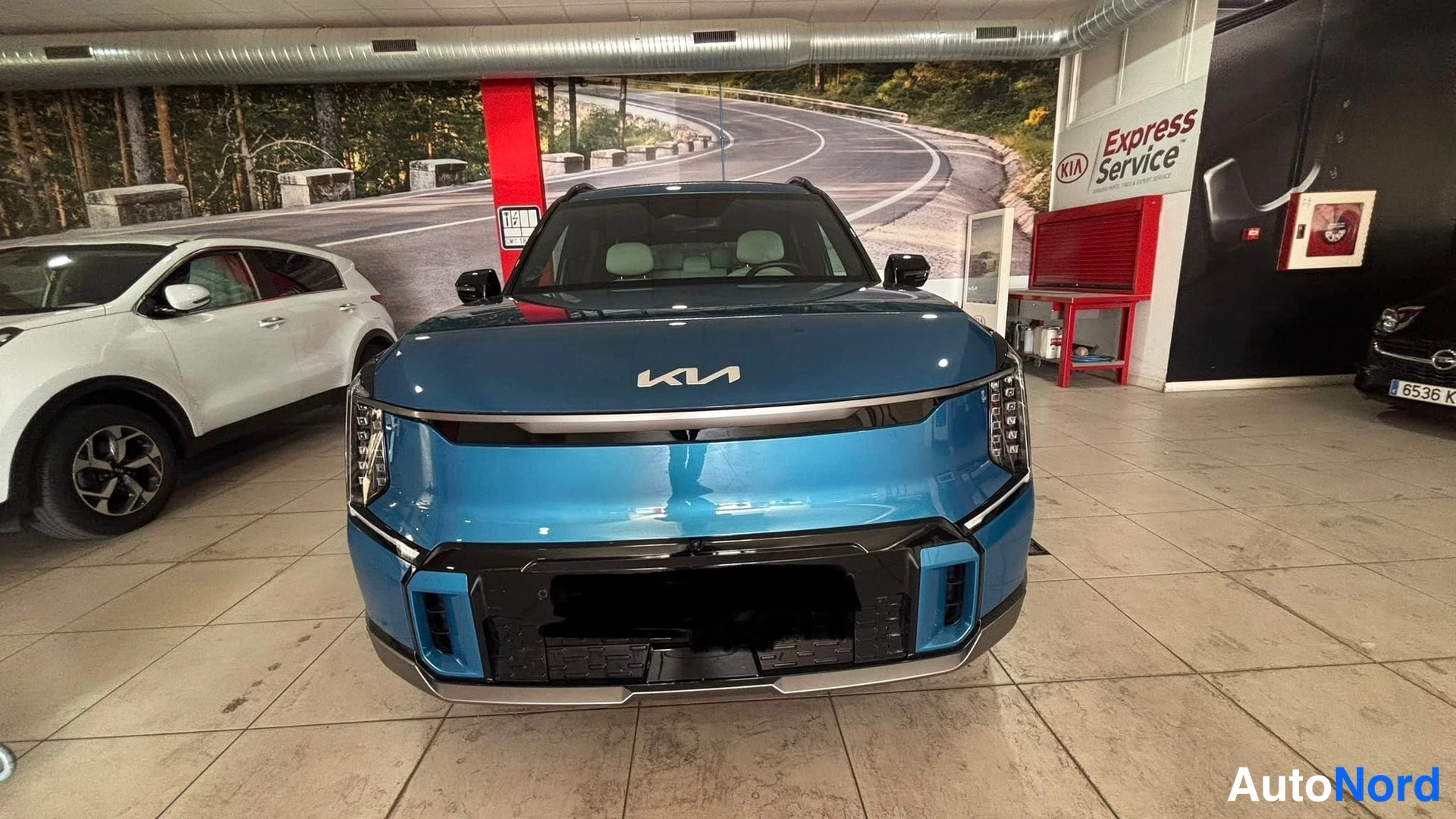 Edestä: Tumansininen Kia-EV9 on suoraan edestä kuvattuna showromssa. Näkyvissä etusuoja, LED-valojen pylväät ja Kia-merkki.