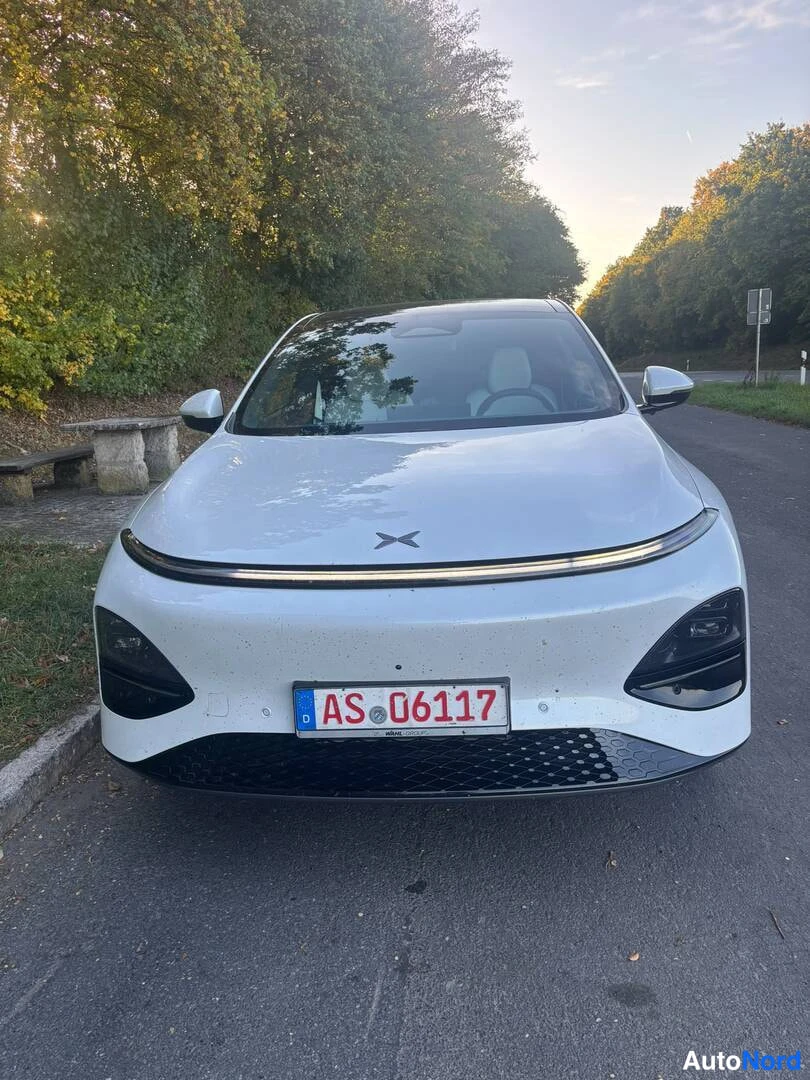 Edestä: Etukulmakuva valkoisesta autosta: etusäleikkö, ohut LED-päävalouhi ja Eurooppa-rekisterikilpi AS 06117 näkyvät puiden reunustamalla tiellä.