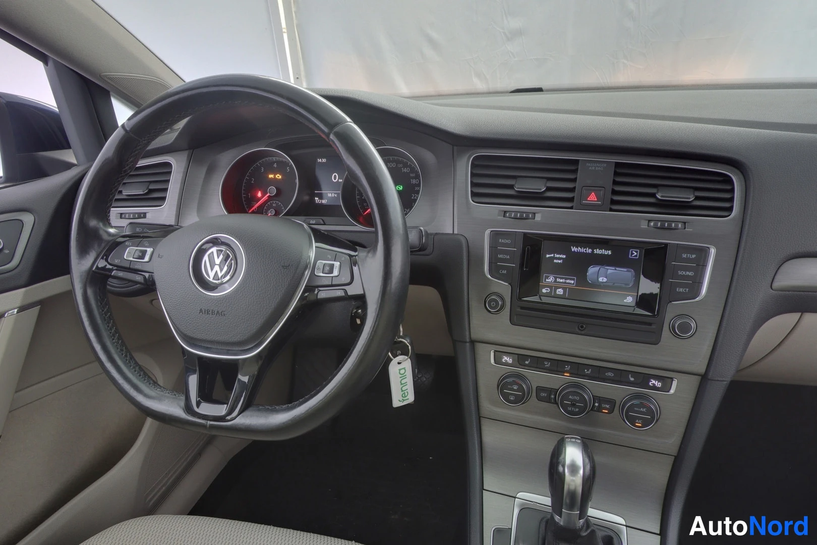 Ohjaamo: Close-up of driver area: steering wheel with multifunction controls, instrument cluster with illuminated warning lamp, centre console touchscreen displaying Veh