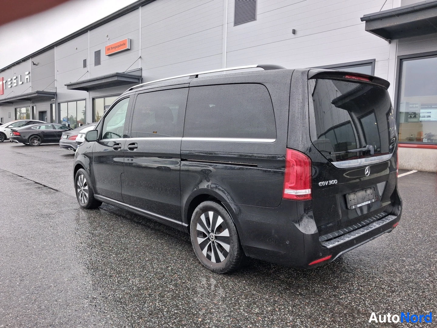 Takaviisto: Mustan Mercedes-Benz EQV:n takaviisto vasemmalta, taa-ikkuna ja punaiset takavalot, autoliikkeen rakennuksen edessä märällä asfaltilla.