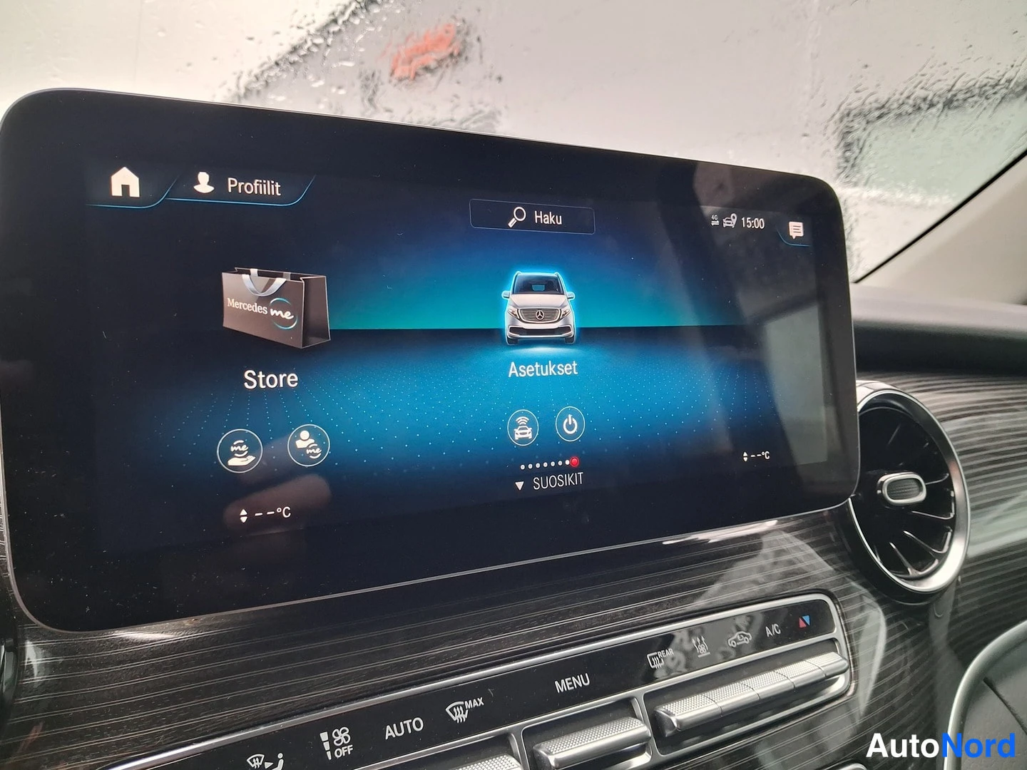 Sivulta: Mercedes-Benzin infotainment-näytön etusivu: valikossa näkyvät Profiilit, Haku, Store ja Asetukset, keskellä MBUX-grafiikka ajoneuvosta; alapuolella kiinteät il