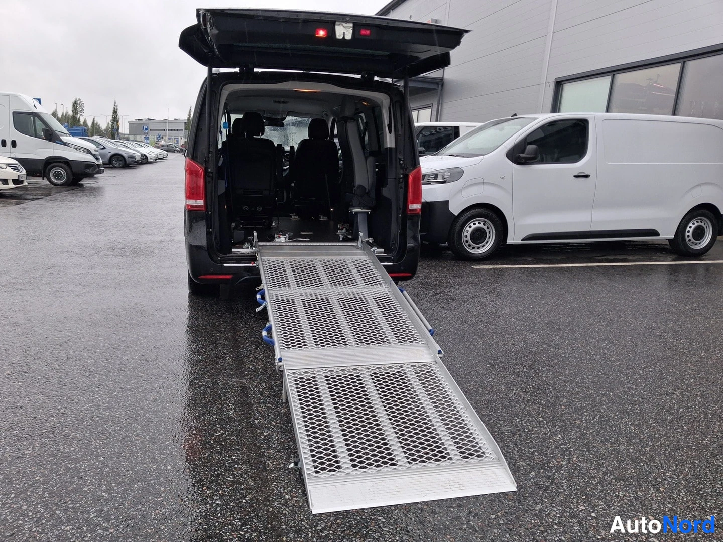 Takaa: Mustan pakettiauton takaosa avoimella takaluukulla ja alumiinisella pyöränosturampilla takaovista kohti maata.