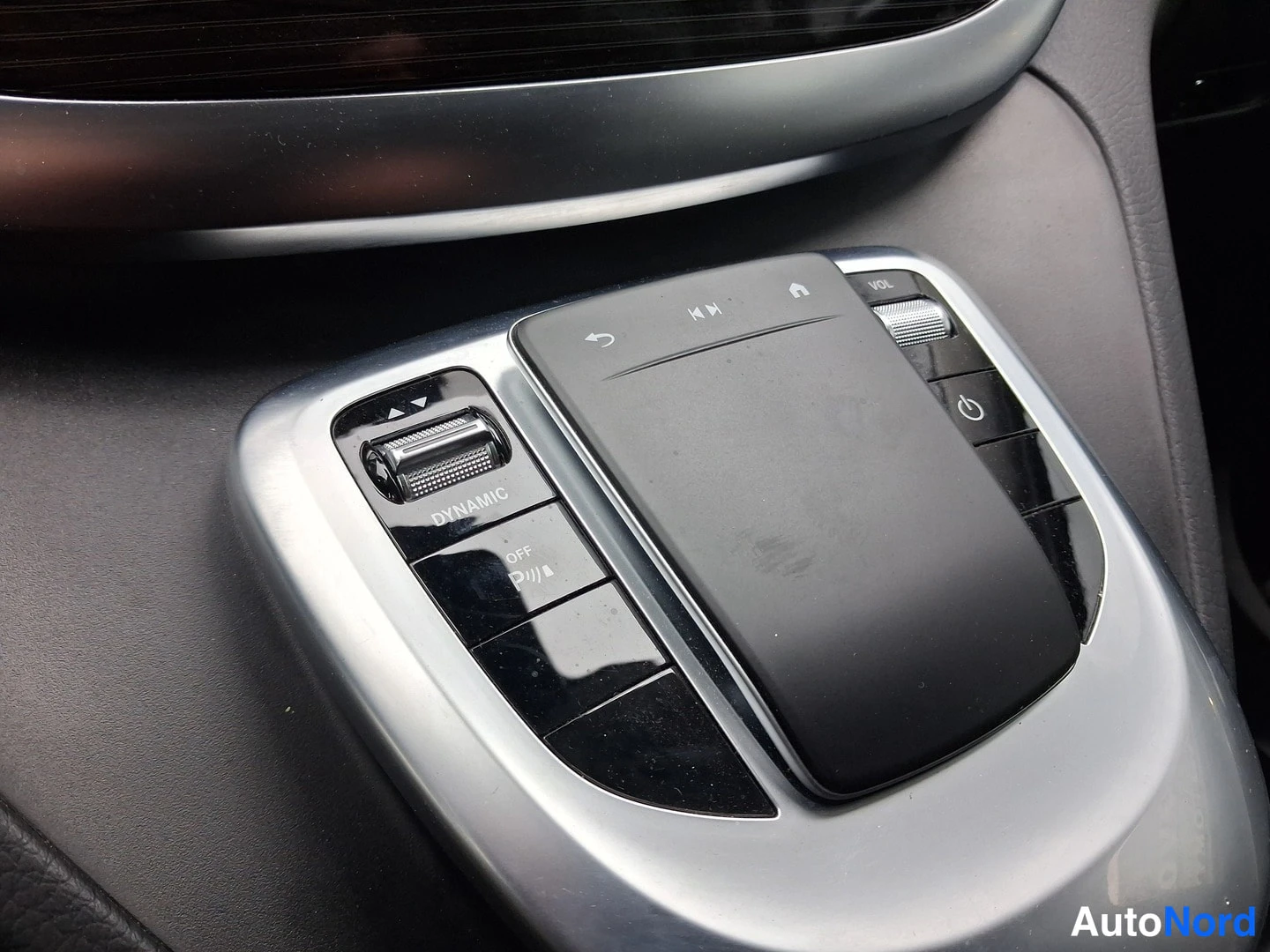 Yksityiskohta: Close-up of the center console controller panel. Features a central touchpad, a 'DYNAMIC' mode button with selector arrows to its left, and volume and power but