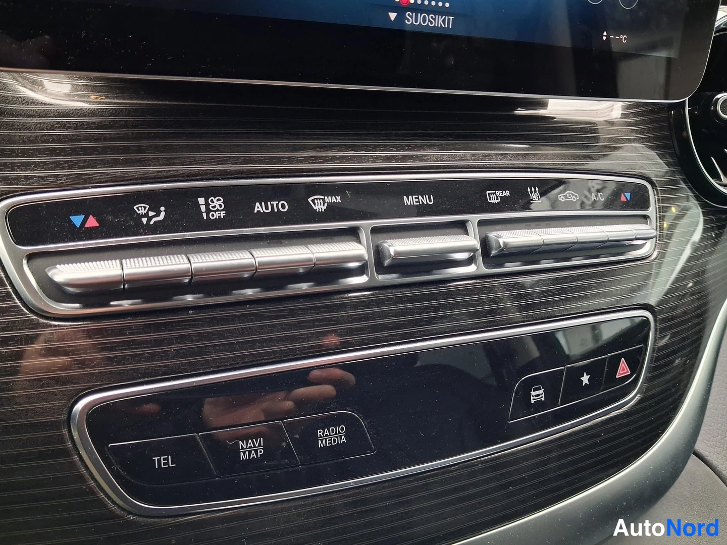 Yksityiskohta: Dashboard climate control interface featuring three metallic slotted rollers. Above are icons for temperature adjustment (red/blue triangles), fan control with