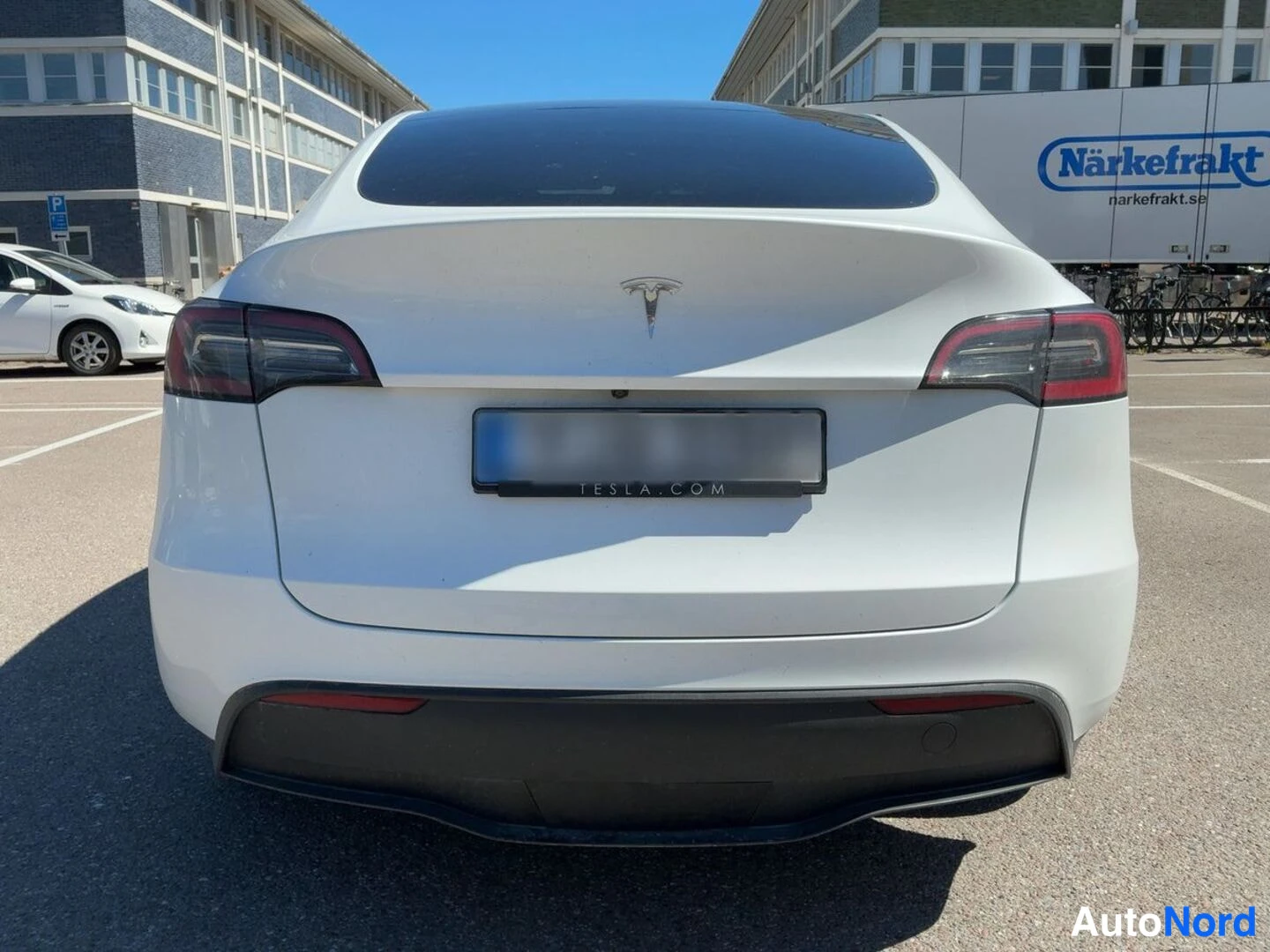 Takaa: Valkoinen Tesla Model Y suoraan takaa kuvattuna; takavalot, T-merkki ja TESLA.COM-tekstiä kantava rekisterikilventeline näkyvissä.