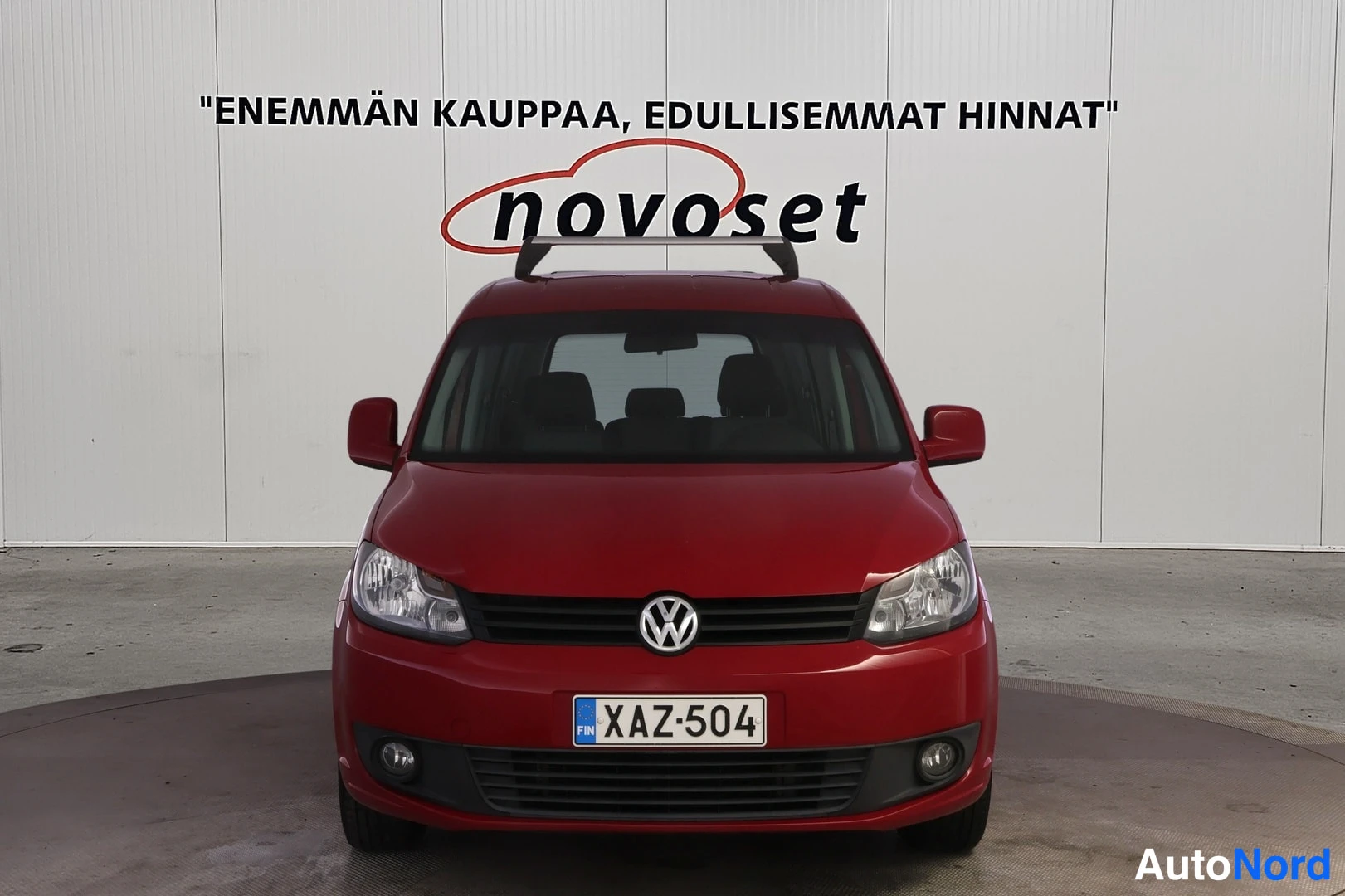 Edestä: Front view of a red Volkswagen with license plate visible on the front bumper. White wall behind vehicle shows Novoset branding. Front grille, headlights, and V