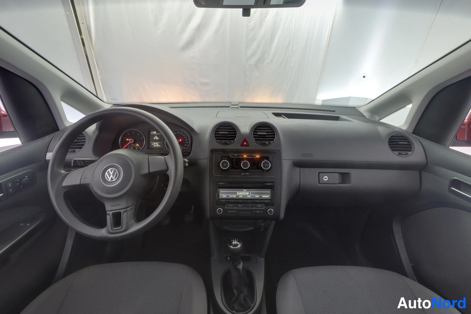 Ohjaamo: Interior cabin view showing dashboard with climate controls, radio unit and manual gear stick steering wheel with VW logo instrument panel behind. Gray fabric s