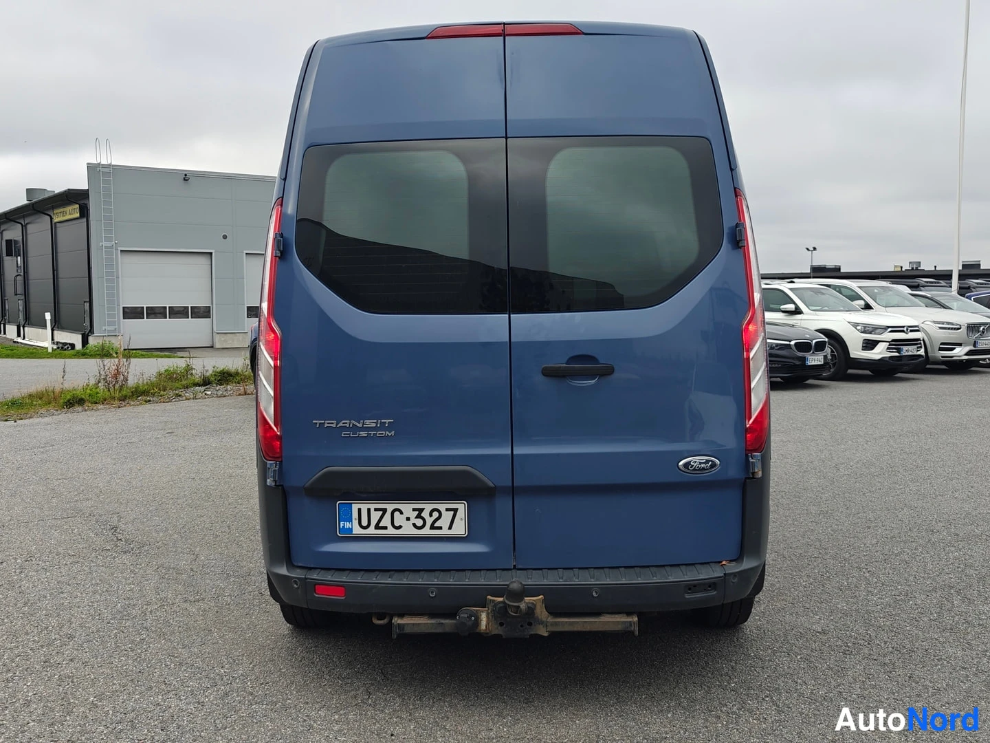 Takaa: Ford Transit Customin takaosa suoraan takaa, takaluukun TRANSIT CUSTOM -teksti, -kilpi ja kytkinlaite puskurin alla.