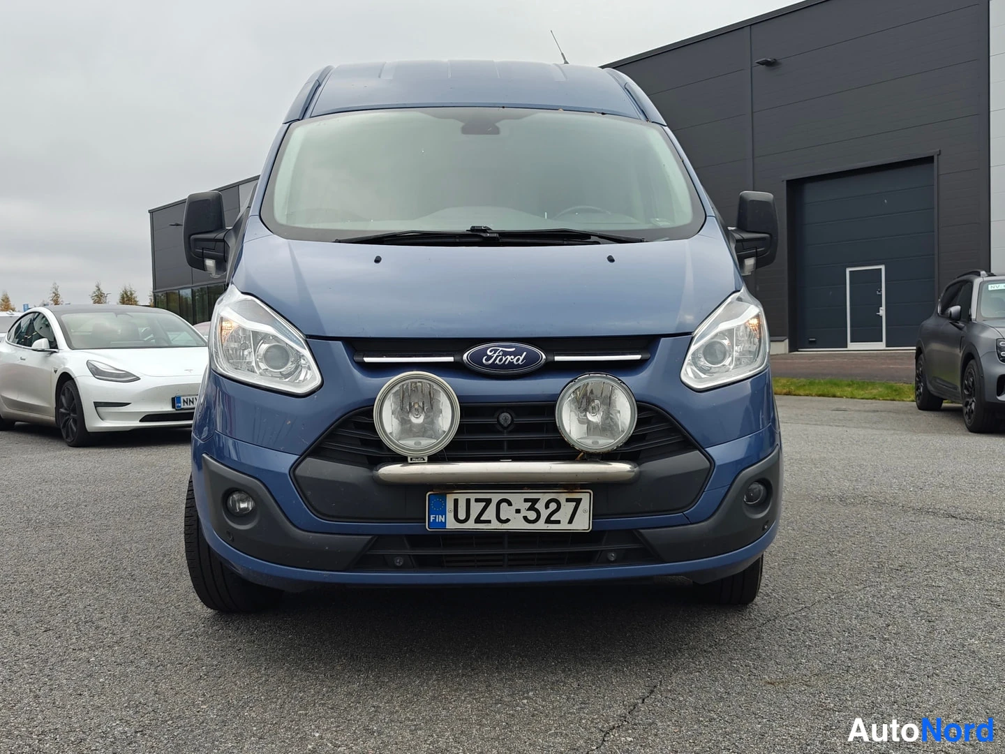 Edestä: Sininen pakettiauto suoraan edestä kuvattuna; Ford-merkki keulassa, kaksi pyöreää lisävaloa puskurissa ja rekisterikilpi.