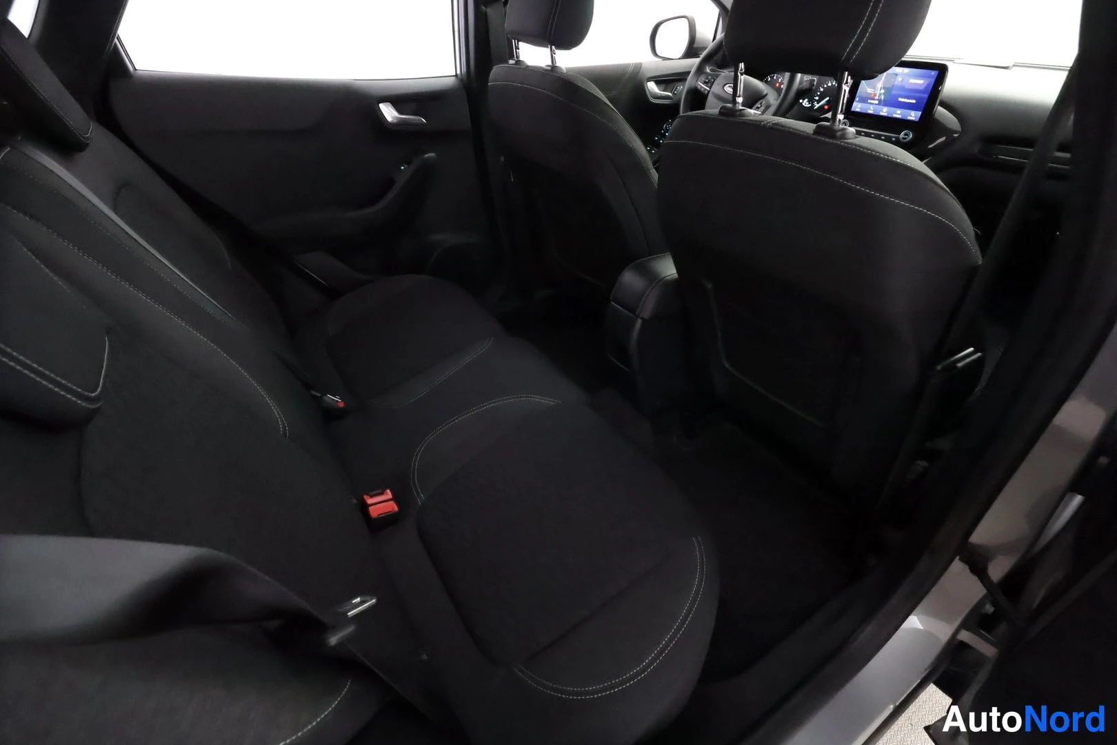 Takaistuimet: Rear seat area showing the black bench seat and headrests. Red seatbelt buckles are attached to the seat base. The back of the front passenger seat and part of