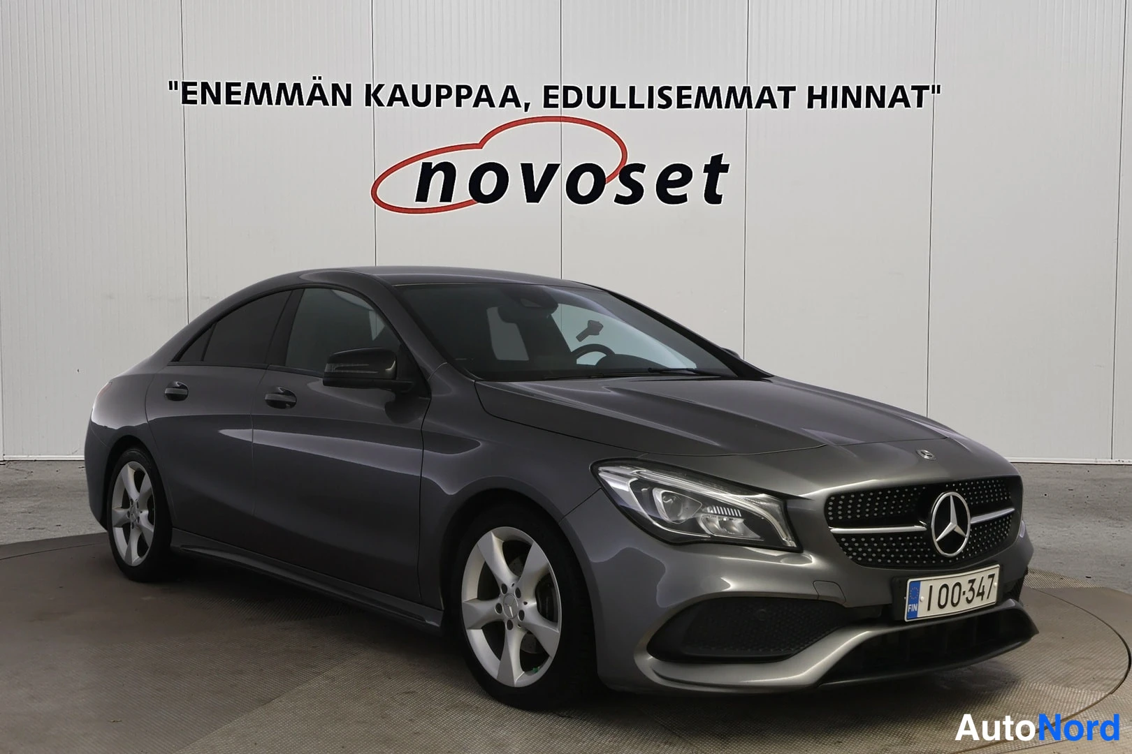 Etuviisto, nokka oikealle: Harmaa Mercedes-Benz CLA -coupe kolmivärikulmassa etunohta ja vasenta kylkeä näkyen, valkoinen Novoset-mainostaulu taustalla, rekisteritunnus 100-347.
