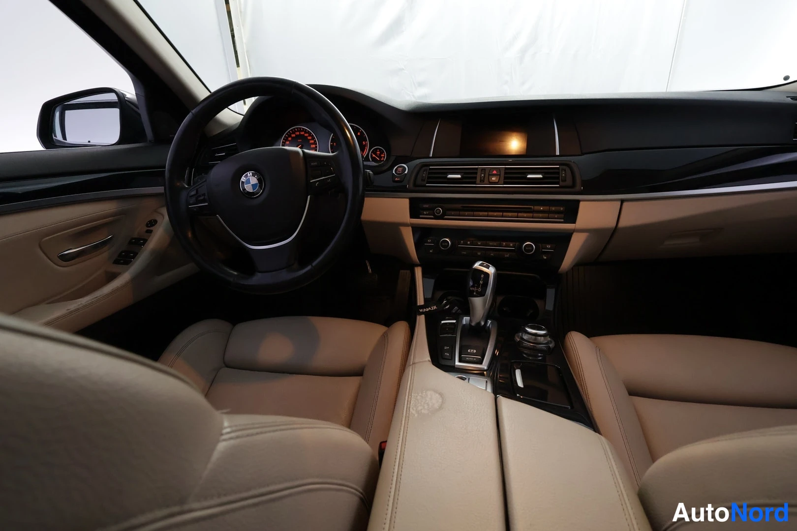 Muu: Kuljettajan puolen sisätila BMW-ohjauspyörällä, takaosalta kuvattuna; beige penkit ja tumma kojelauta.