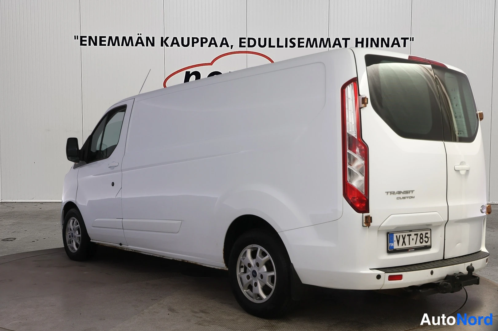 Takaviisto: Valkoinen Ford Transit Custom -pakettiauto takaviistosta; TRANSIT CUSTOM -merkintä, takavalot ja rekisteritunnus näkyvät.