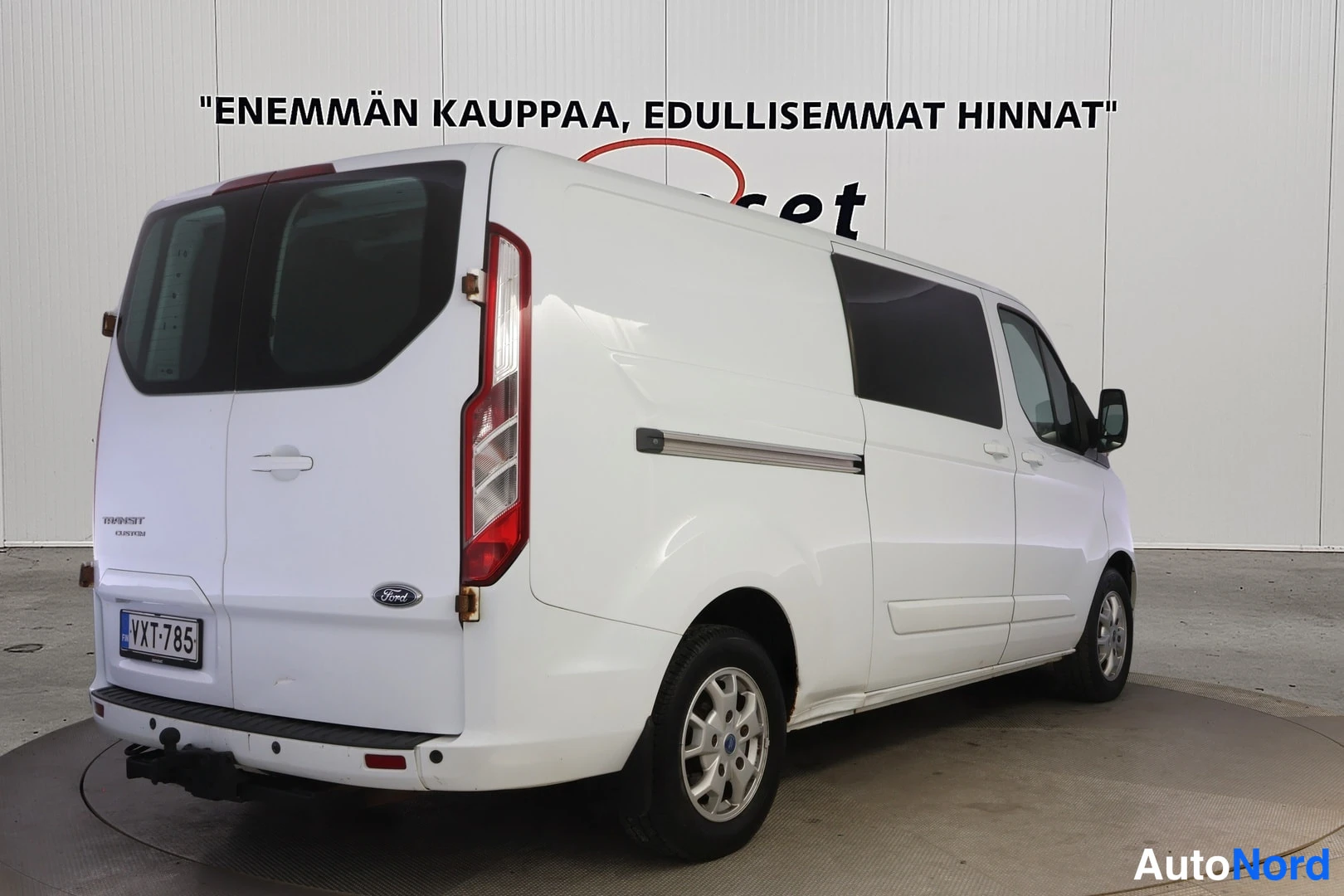Takaviisto: Valkoinen Ford Transit Custom -pakettiauto takaviistosta toiselta sivulta; takavalot, rekisteritunnus ja vetokoukku näkyvät.