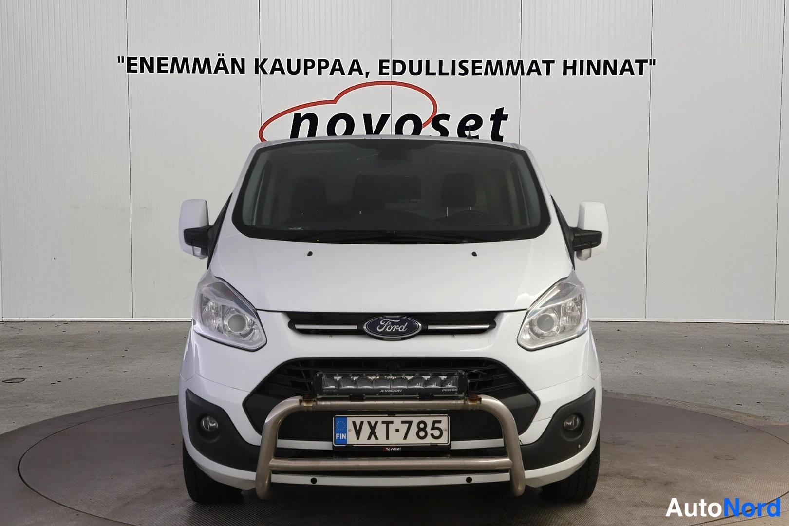 Edestä: Ford Transit Custom -pakettiauto suoraan edestä kuvattuna: valkoinen kori, Ford-merkki keulassa, puskurikaari ja LED-valopalkki, rekisterikilpi.