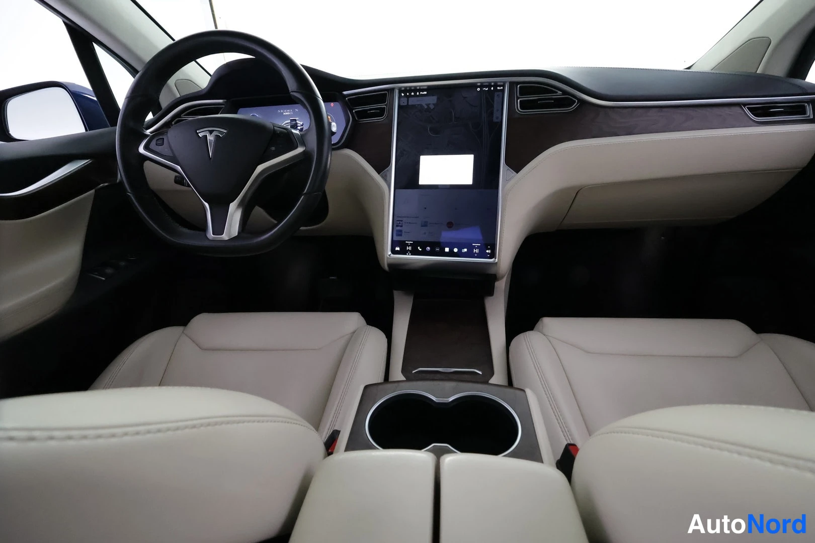 Muu: Täysin suunnattu kuva kojelaudasta: Tesla-merkitty ohjauspyörä, keskellä suuri pystynäyttö, vaaleat etuistuimet ja keskikonsolin kupiteline.