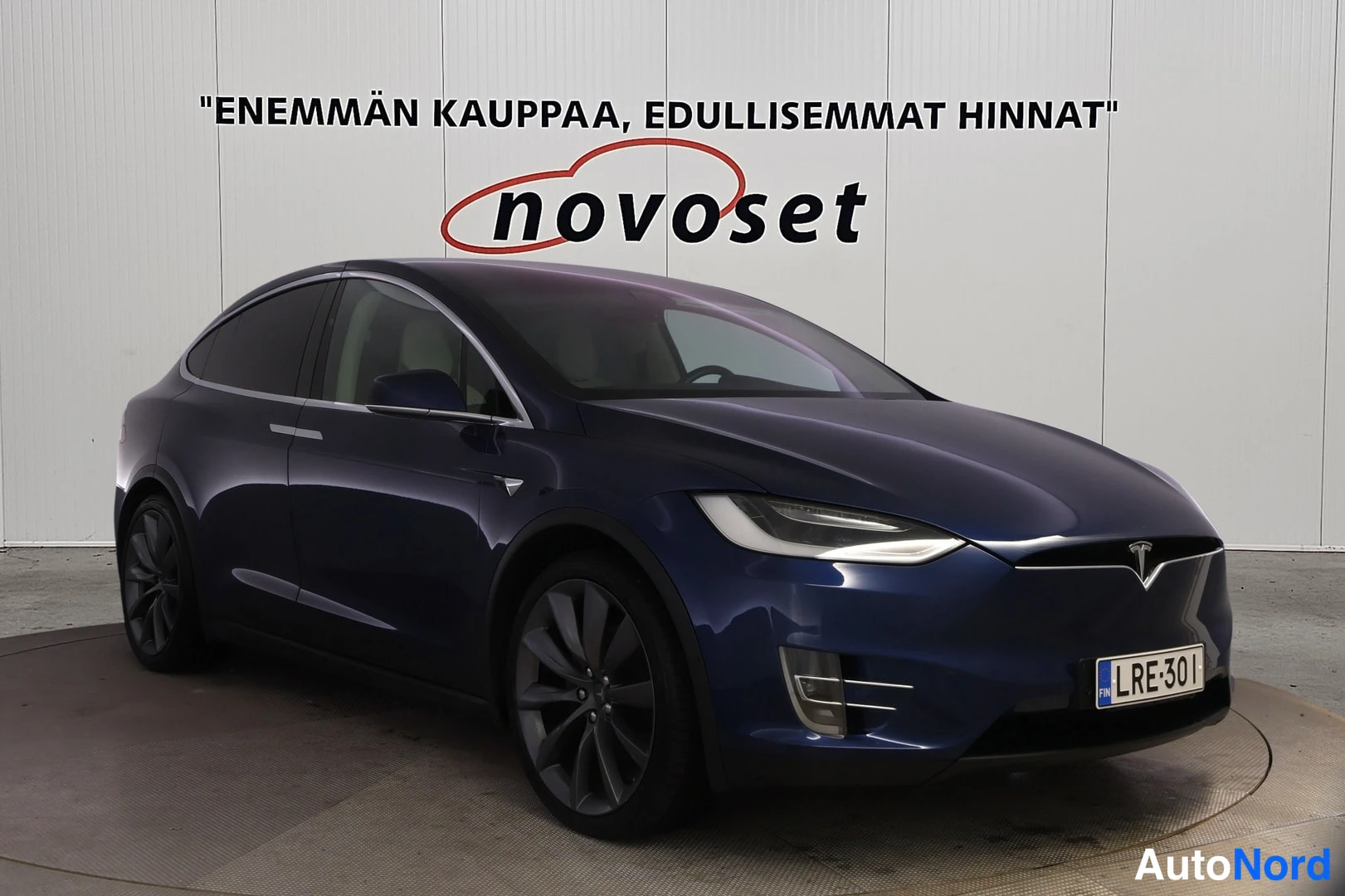 Etuviisto, nokka oikealle: Tummansininen Tesla Model X etukolmannesnäkymässä, keula kuvan oikeassa reunassa; näkyvissä ajovalot, etusäleikkö, rekisterikilpi ja tummat kevytmetalliv