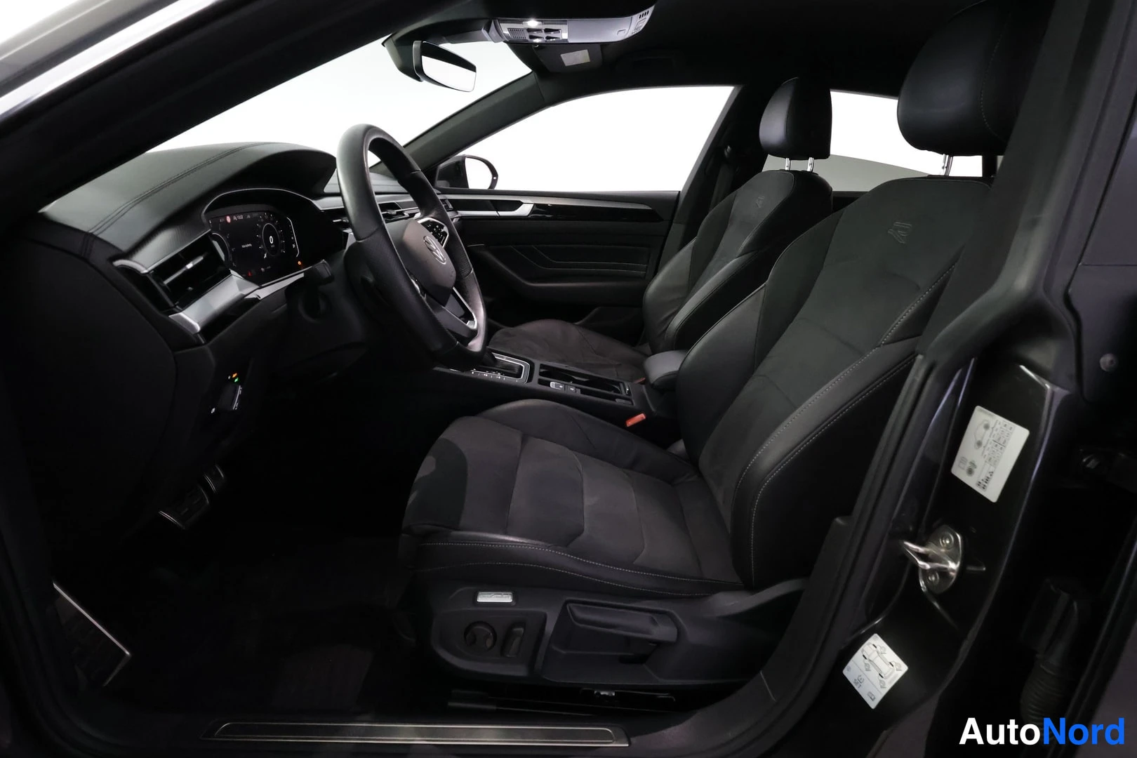 Ohjaamo: Interior shot looking through the open driver's door. The two black front seats are in focus, flanking the center console. The steering wheel and dashboard curv