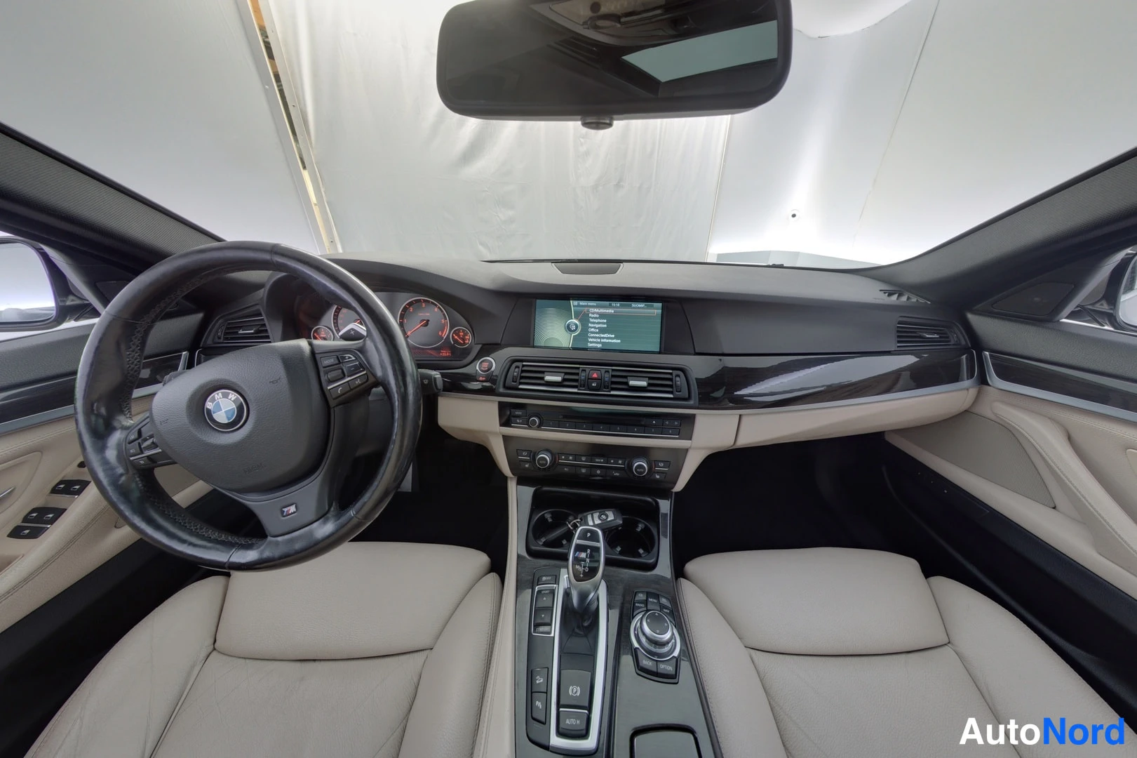 Muu: Näkymä ohjaamoon takaa eteenpäin: BMW-ohjauspyörä, kojetaulu ja keskonitori, jonka valikossa teksti COMMUNICATION, automaattivaihteen valitsin ja beiget istuime