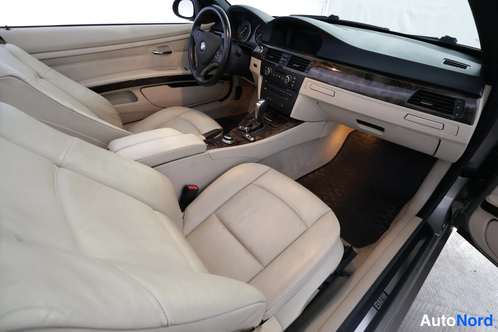 Yksityiskohta: Etumatkustamo ohjauspyörällä, mittaristolla ja keskikonsolilla, beige nahkaverhoilu ja BMW-kynnyksellä.