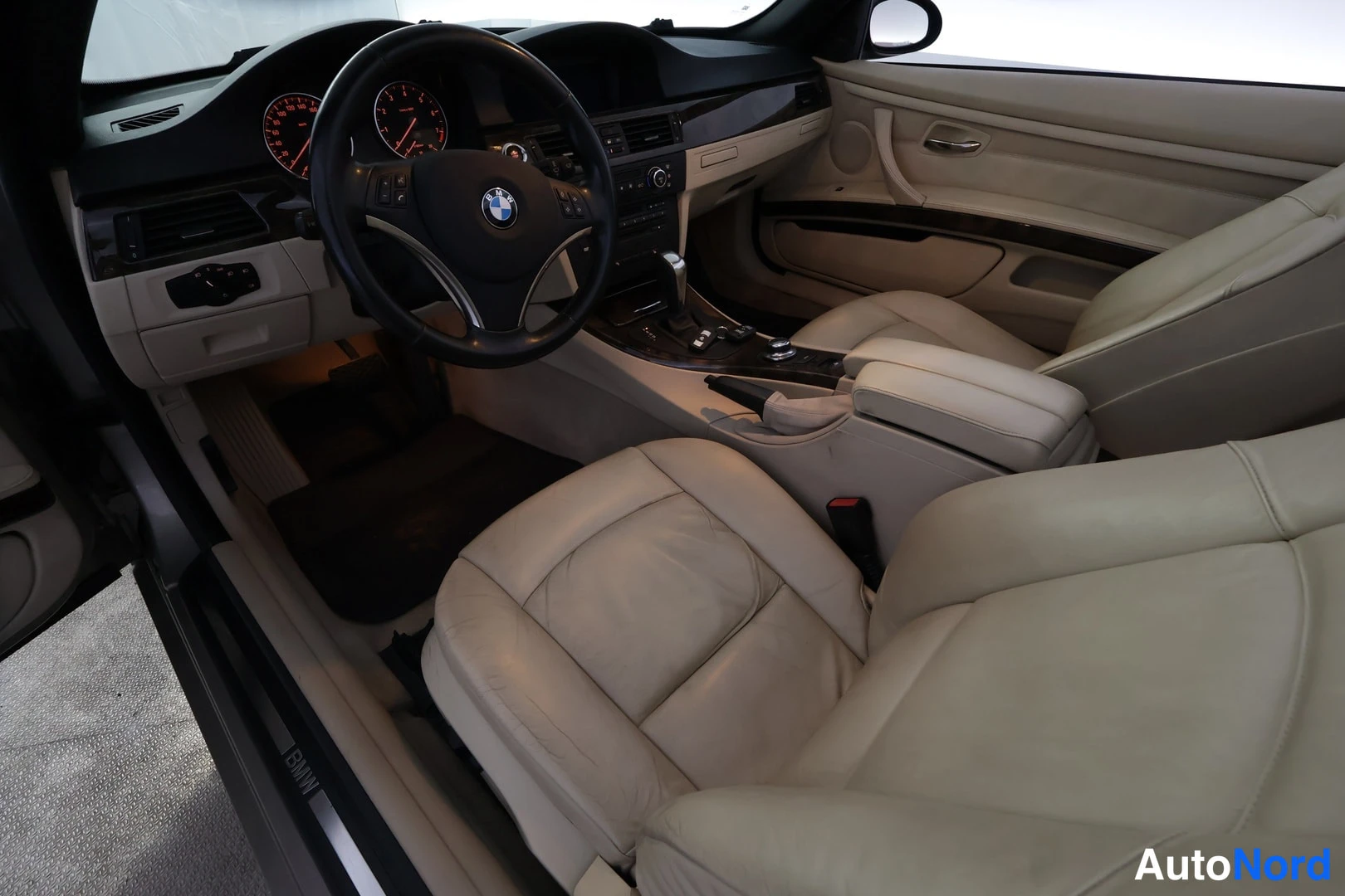 Ohjaamo: Ohjaamokuva: ohjauspyörä BMW-logolla, mittaristo punaisilla viivoilla, keskuspaneeli ja etusuodatin tuolien edessä. Ovet ovat auki.