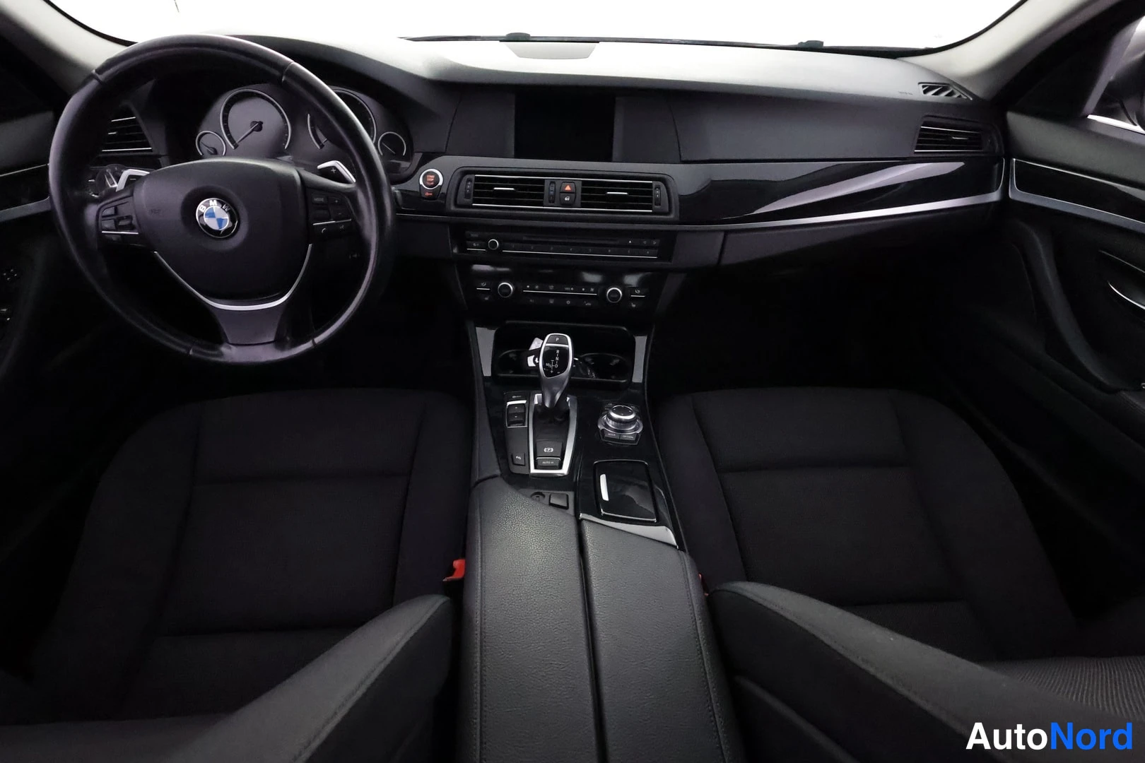 Edestä: Ohjaamo etupuolelta: BMW-ohjauspyörä, mittaristo, keskikonsoli ja vaihdevipu, mustat kankaanverhoillut etuistuimet.