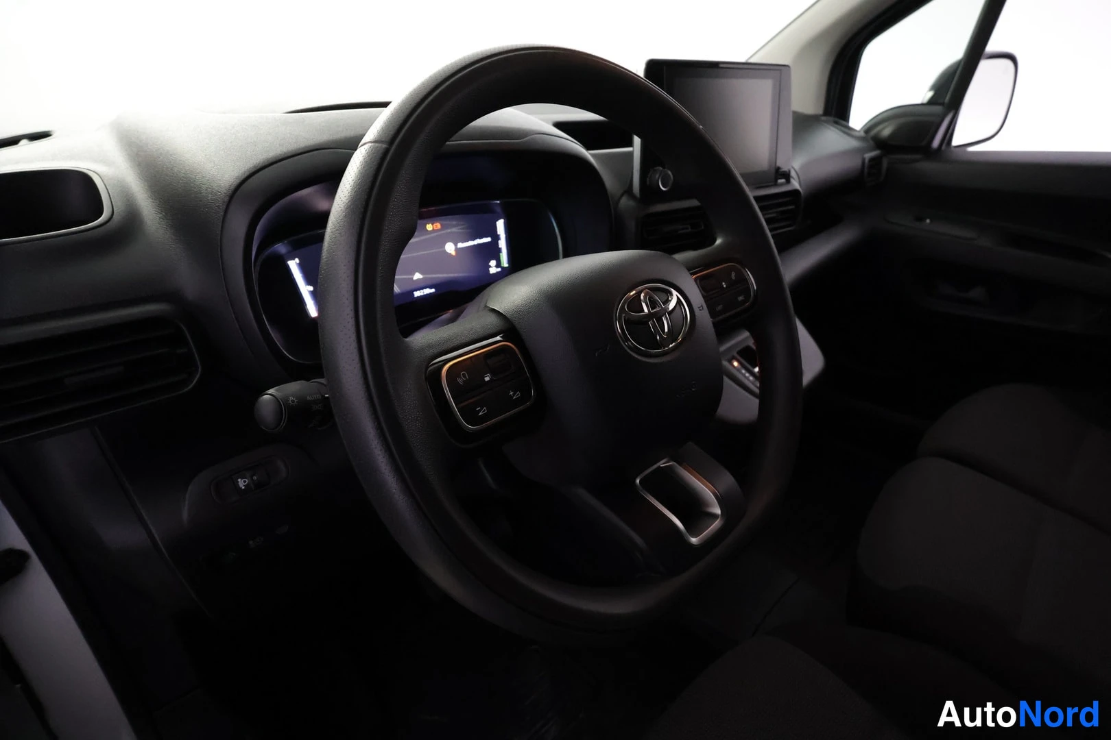 Muu: Auton ohjaamo: musta ohjauspyörä Toyota-merkillä, digitaalinen mittaristo ja keskinäytöllä varustettu kojelauta.
