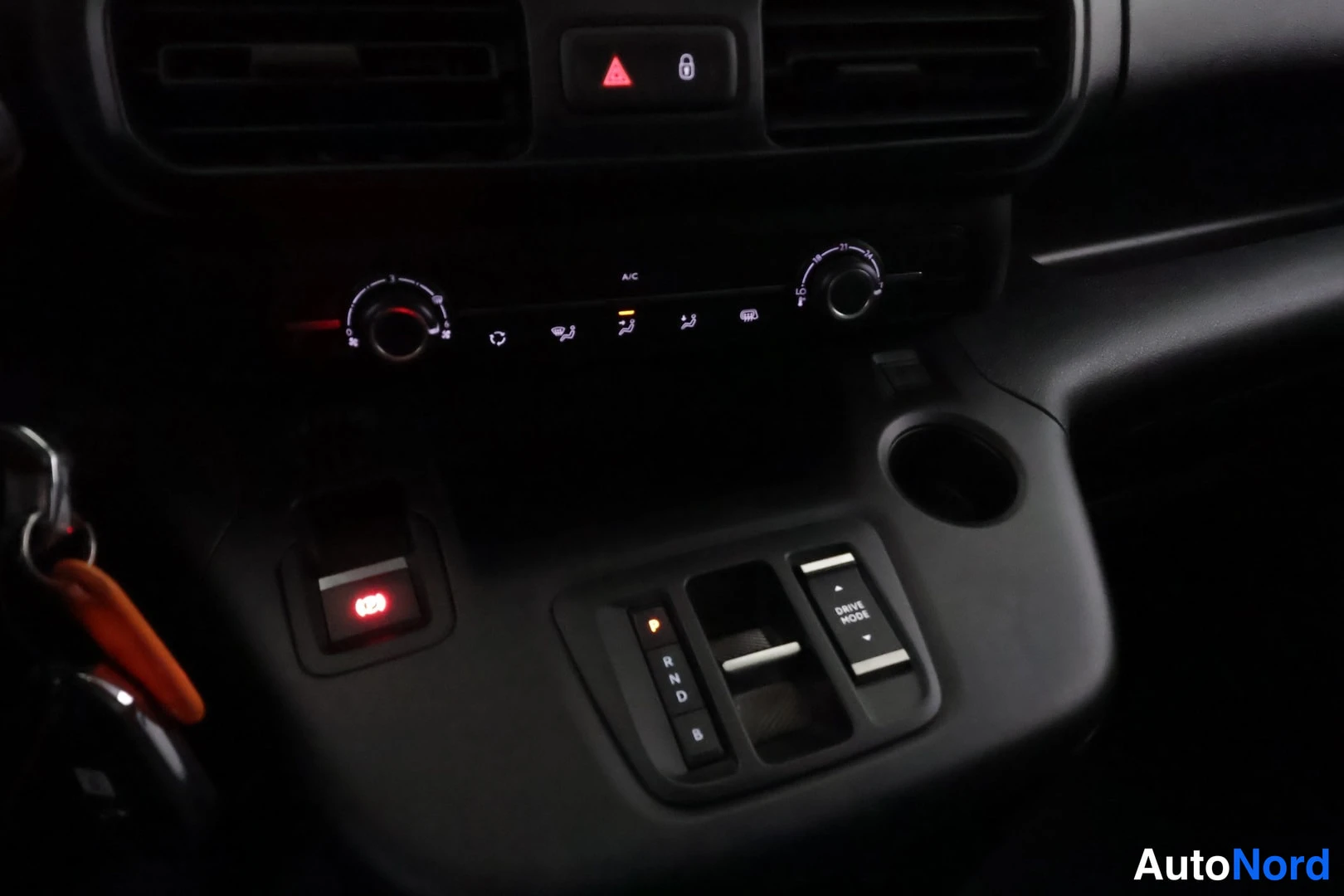Muu: Auton keskikonsoli pimeässä: ilmastointia säätävät kaksi nuppia, A/C-nappi, P R N D B -painikkeet, DRIVE MODE-kytkin ja kuppiteline.