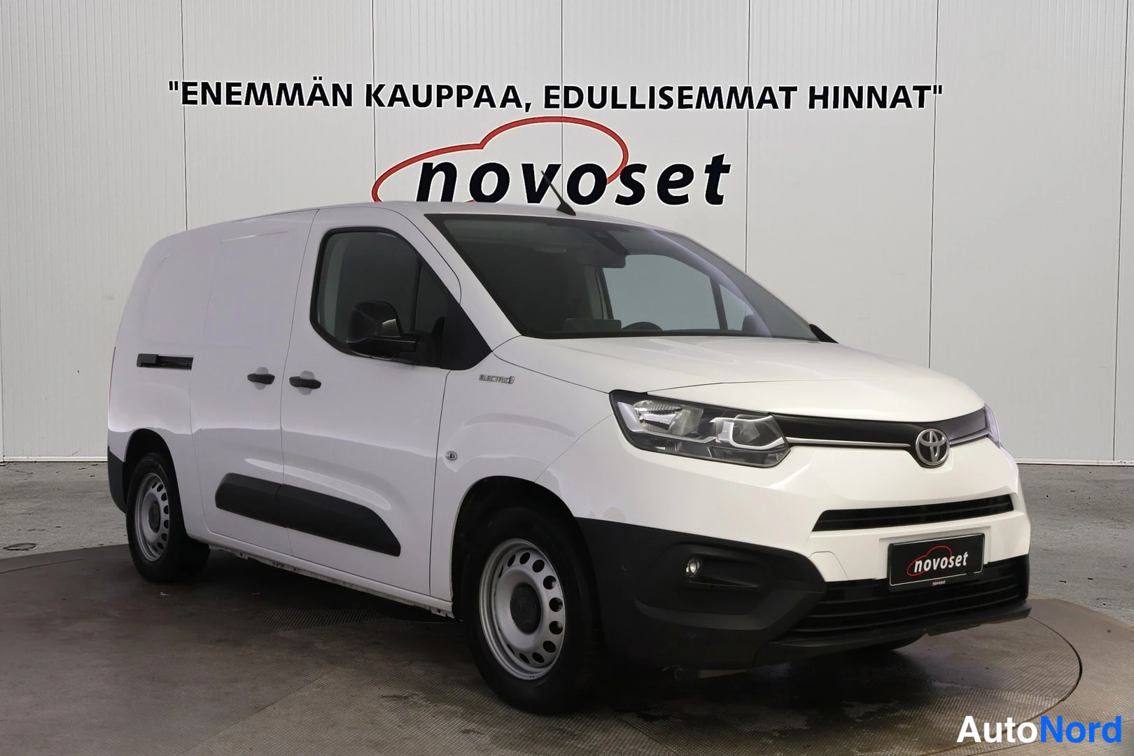 Etuviisto, nokka vasemmalle: Valkoinen Toyota-vanilla ajoneuvo etu- ja sivusta. Ilmoitustaulu takaseinässä.