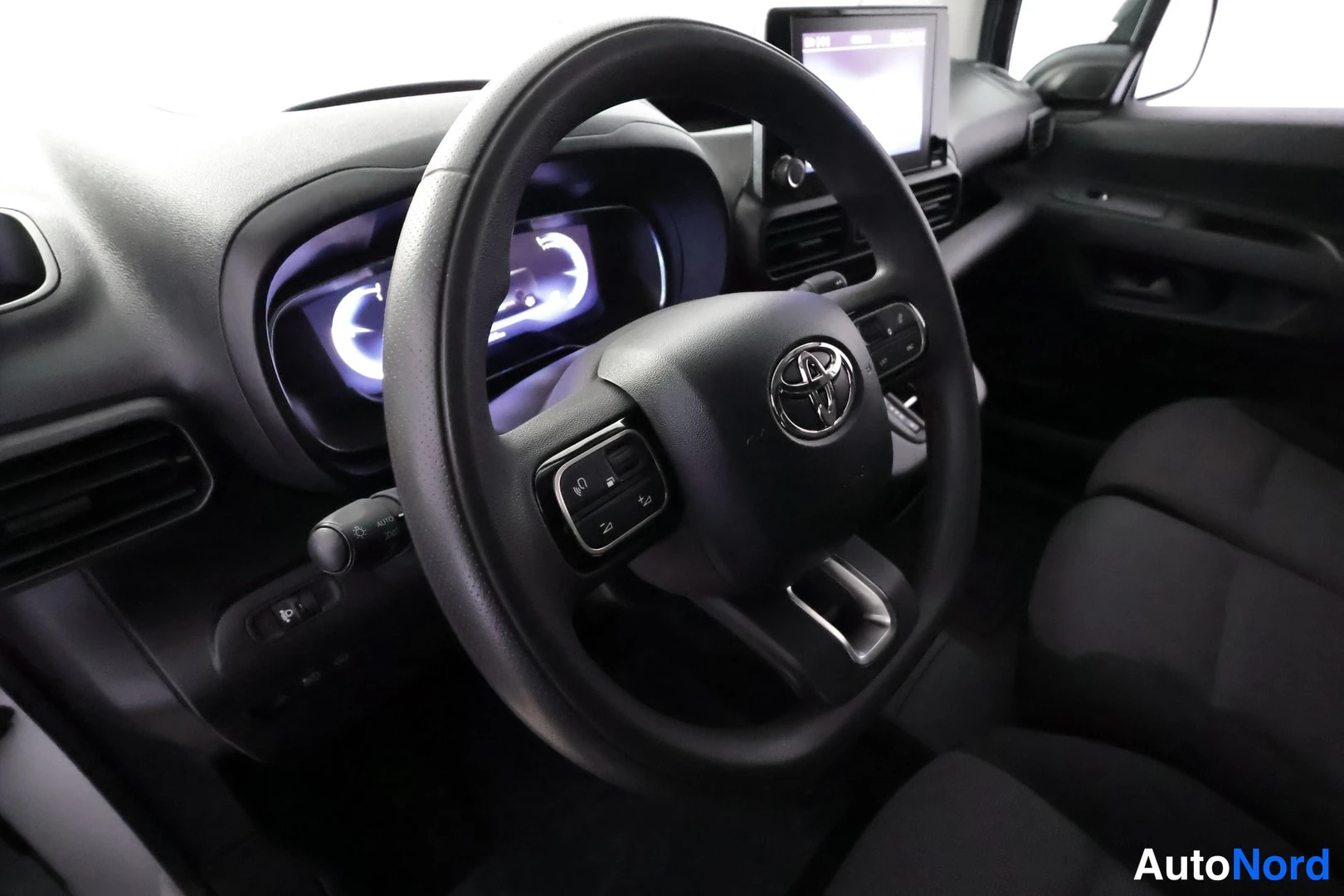 Yksityiskohta: Toyota-merkitty ohjauspyörä, digitaalinen instrumenttinäyttö ja kojelauta keskellä kosketusnäyttö; etupenkit ja tumma sisusta näkyvät.