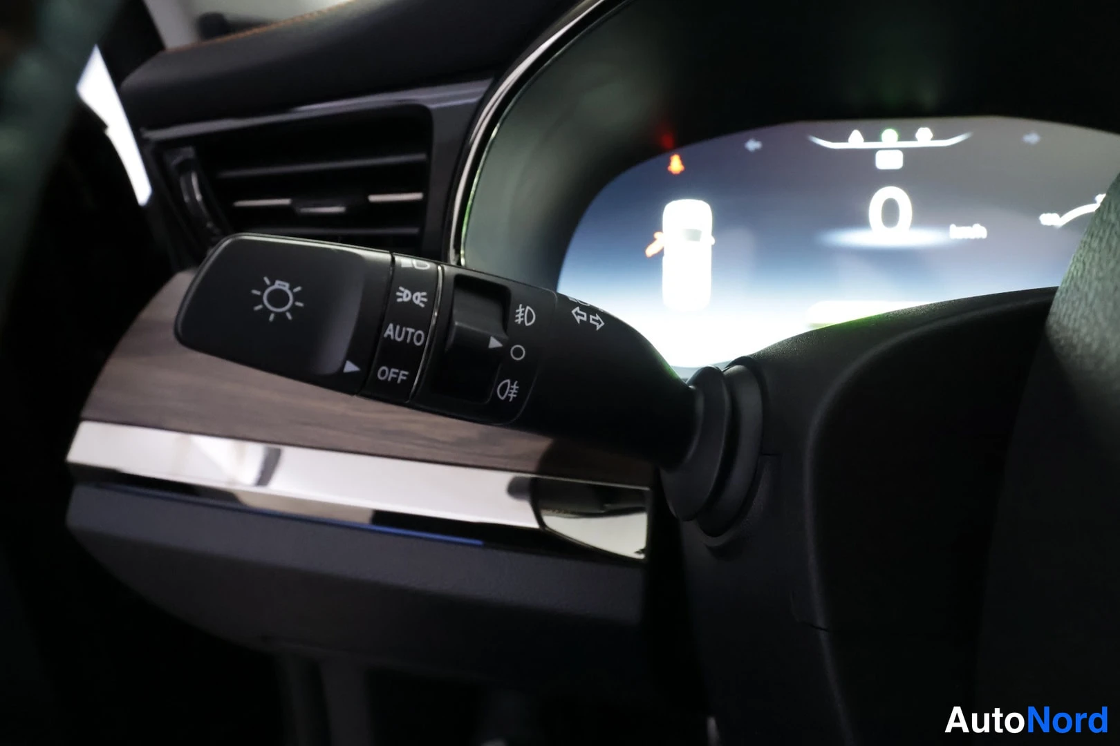 Yksityiskohta: Sisäkuva auton valokytkimestä, jossa on AUTO- ja OFF-asemaa sekä suuntavilkun symboli; kojelaudan näyttö näkyy taustalla.