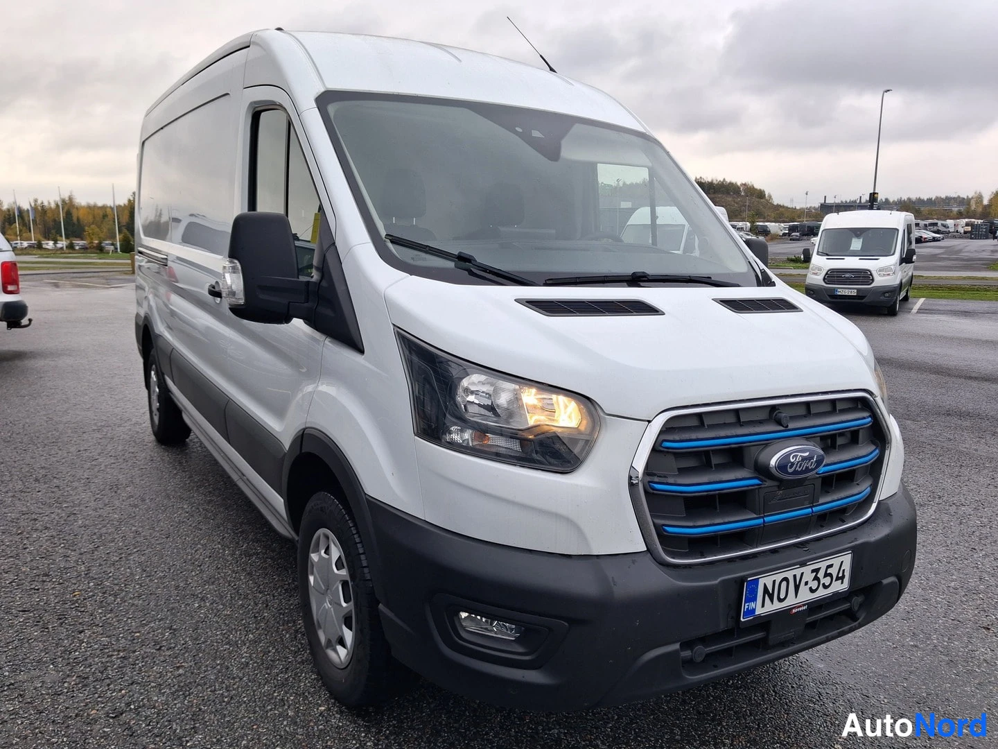 Etuviisto, nokka oikealle: Valkoinen Ford E-Transit-pakettiauto kolmivaiheisesti edestä vasemmalta parkkialueella, tunnus.