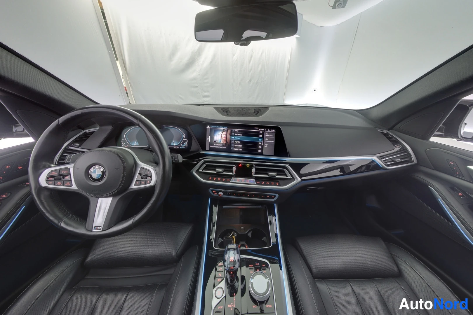 Muu: auton etuistuimilta kuvattu ohjaamo: BMW-ohjauspyörä, mittaristön näytöt ja keskinäytön radiovalikko, mustat istuimet.