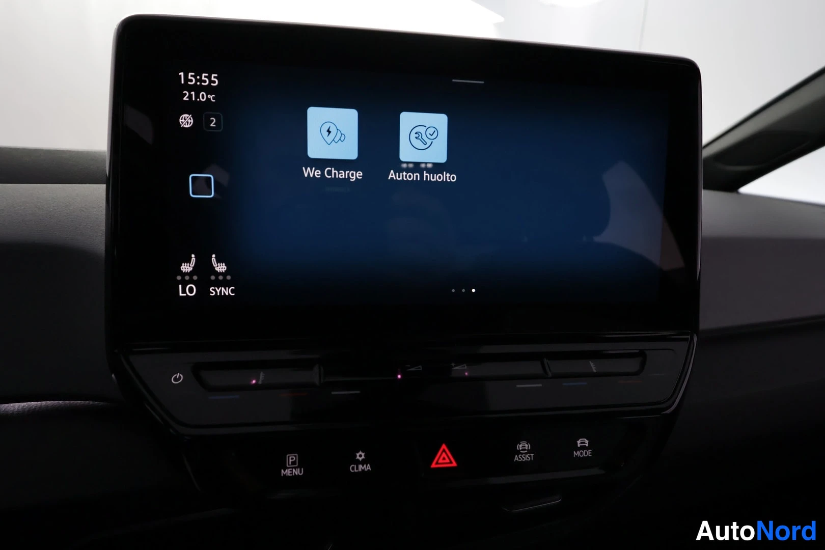 Yksityiskohta: Auton näytöllä We Charge- ja Auton huolto -kuvakkeet, alla kosketuspainikkeet MENU, CLIMA, ASSIST ja MODE.