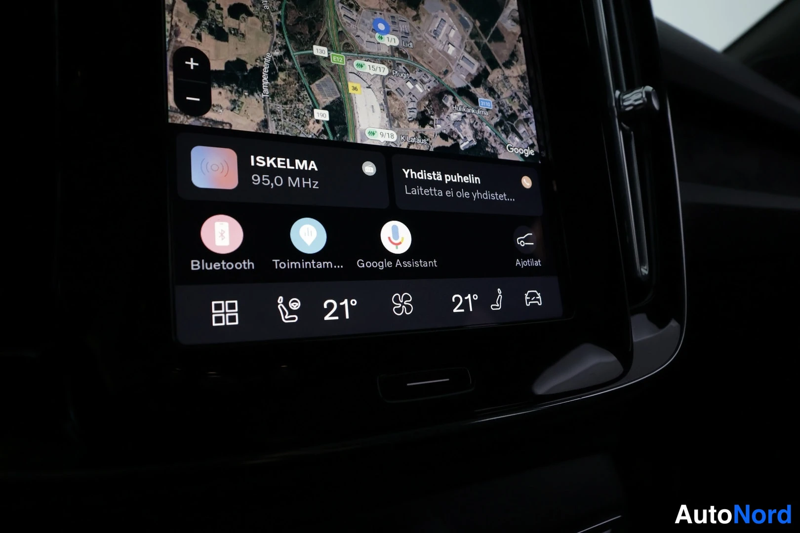 Muu: Infotainment-näytön alaosan lähikuva: ISKELMA-radio, Bluetooth, Google Assistant ja kahden vyöhykkeen lämpötilat 21°.