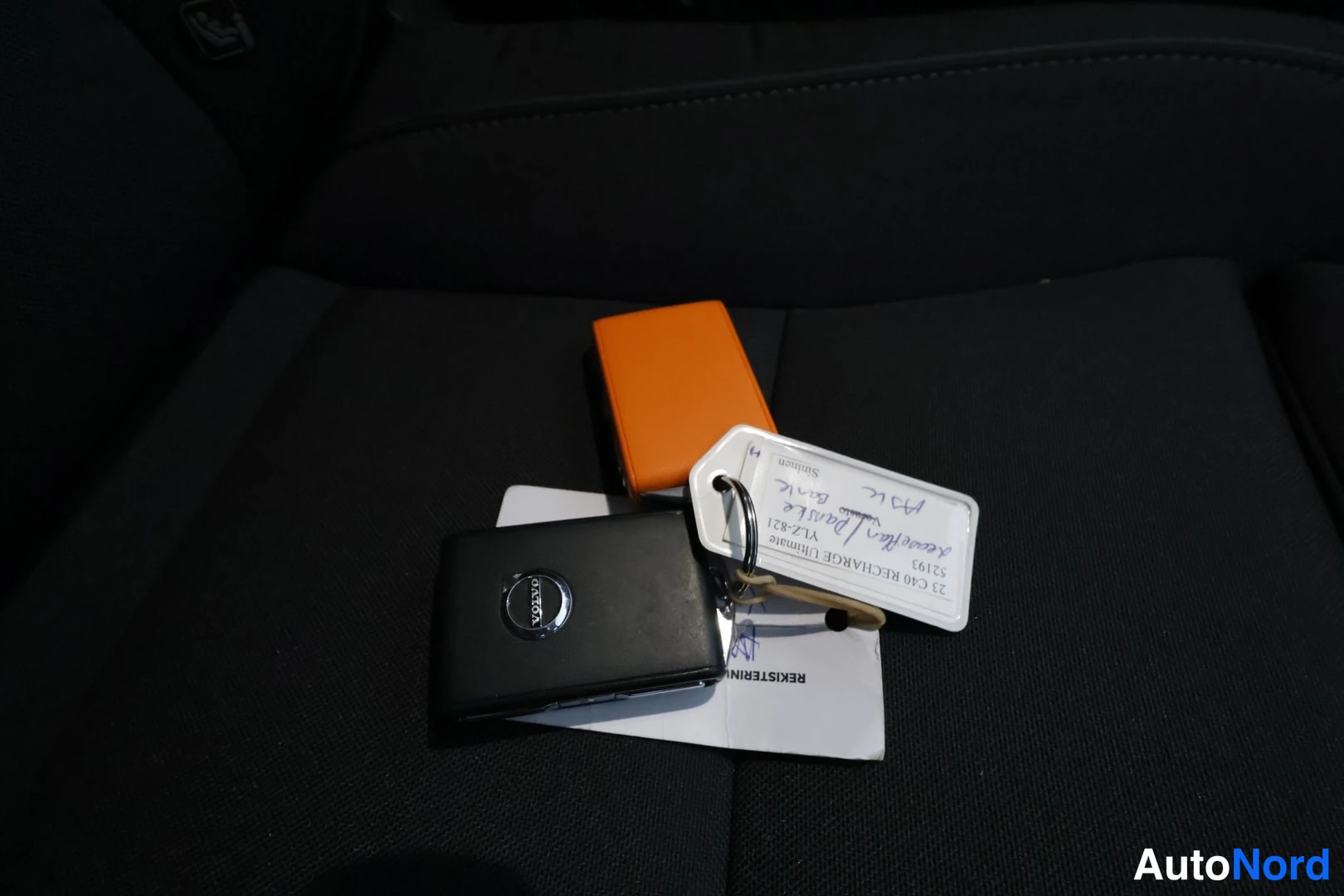 Yksityiskohta: Kuva istuimella makaa musta Volvon avain ympyrälogolla, oranssi avaimenkuori sekä käänteisiä papereita, joissa lukee muun muassa '23 C40 RECHARGE' ja 'REKISTERI