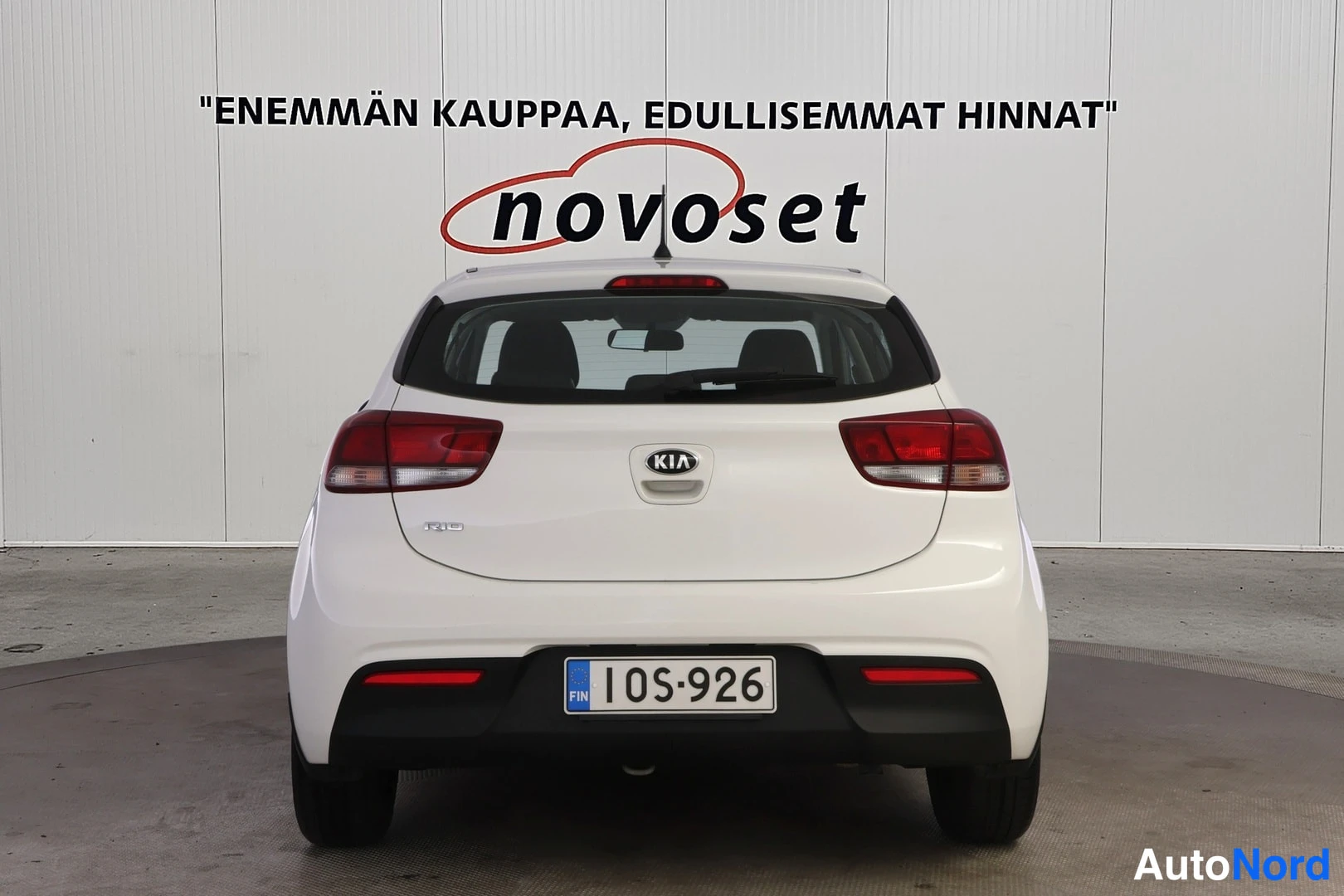 Takaa: Valkoinen Kia Rio -viistoperä suoraan takaa kuvattuna; takavalot, Kia-merkki ja rekisterikilpi 1OS-926 näkyvät.