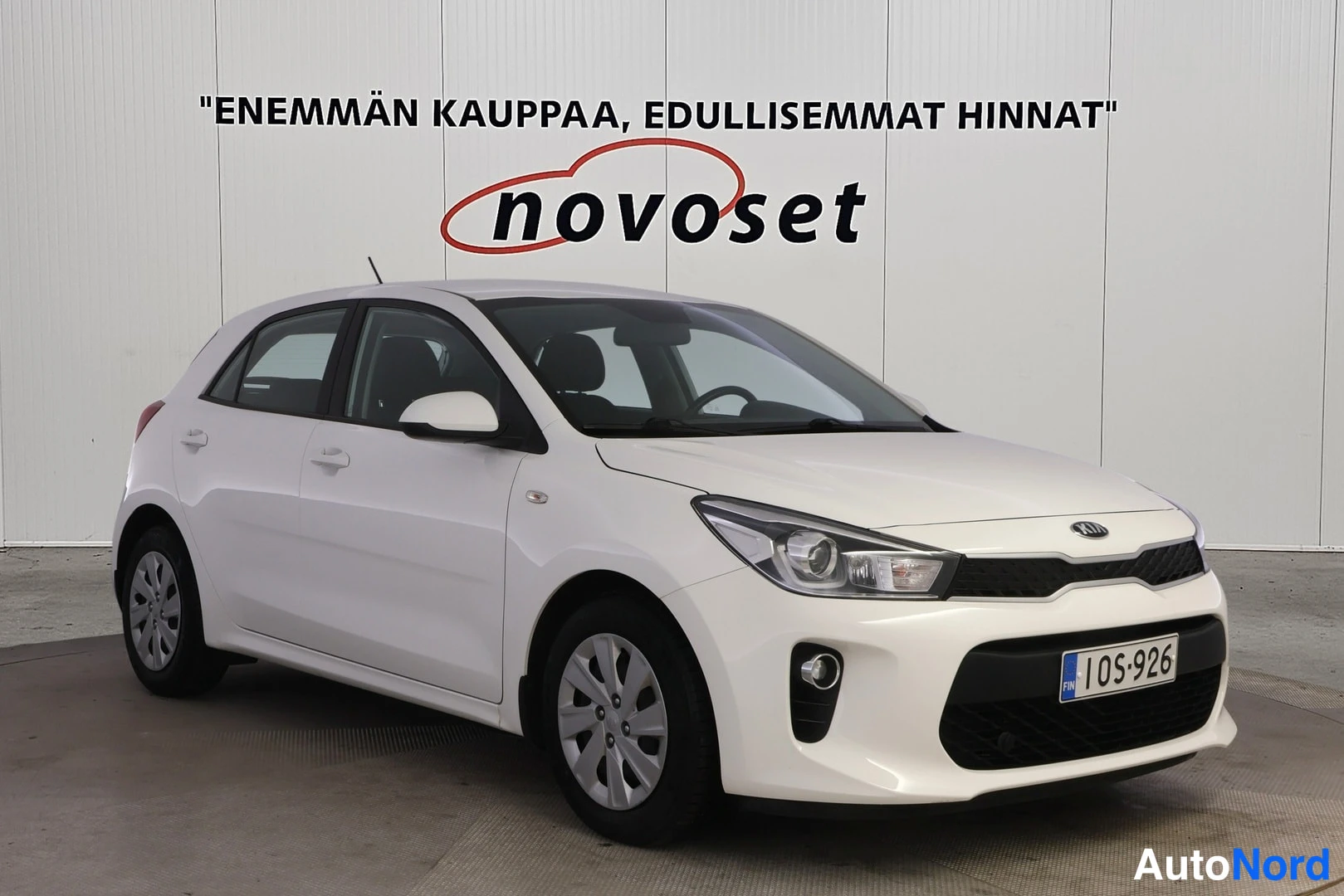 Etuviisto, nokka vasemmalle: Valkoinen Kia-hatchback nähdään edestä ja oikeasta puolelta. Etusarja on musta ja rekisterikilpi lukee 1OS-926. Taustaseinällä teksti novoset.
