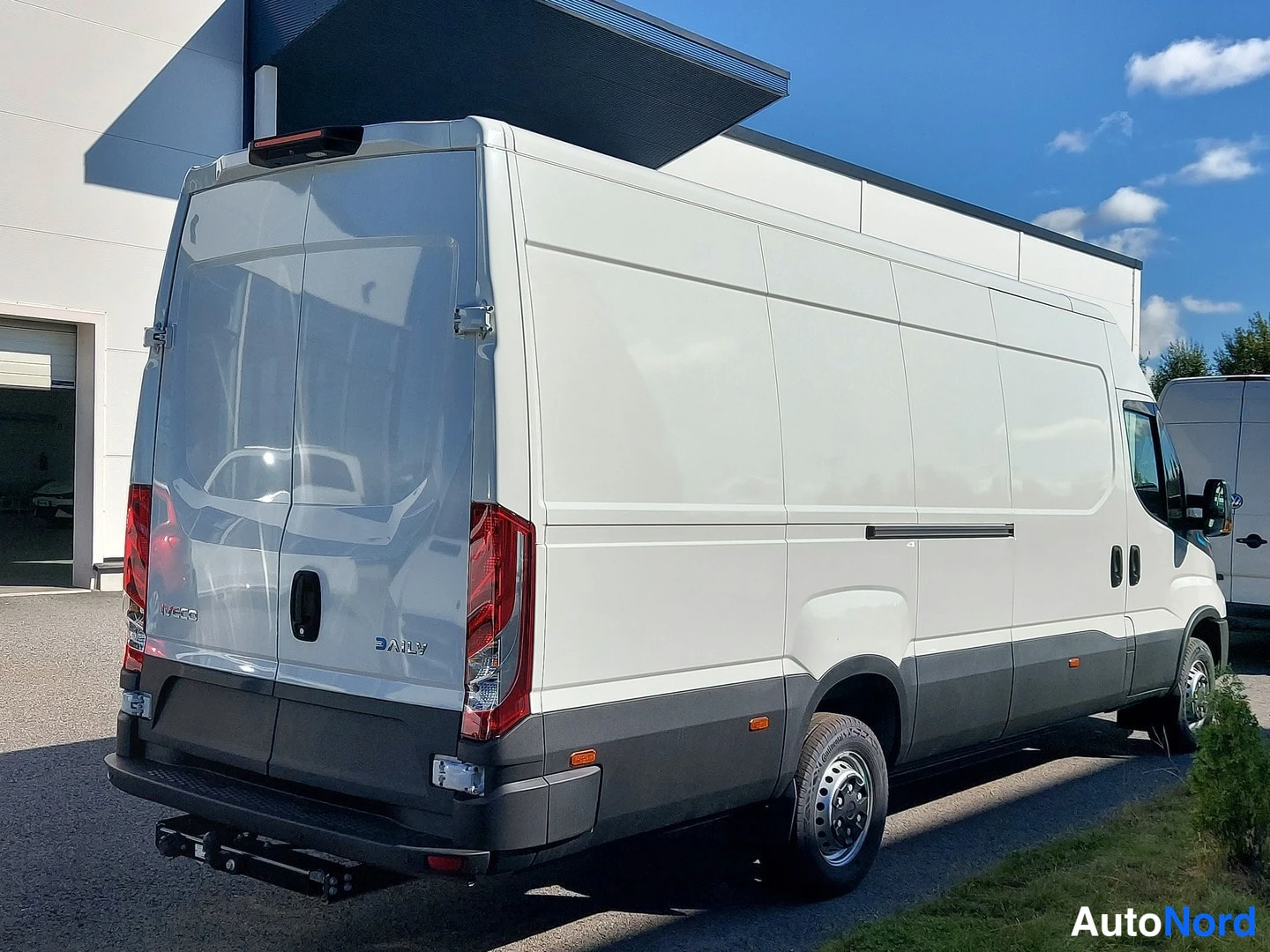 Takaviisto: Valkoinen Iveco Daily -pakettiauto takaviistosta, perä vasemmalla; punaiset takavalot, DAILY-teksti takaluukussa ja vetokoukku takana.