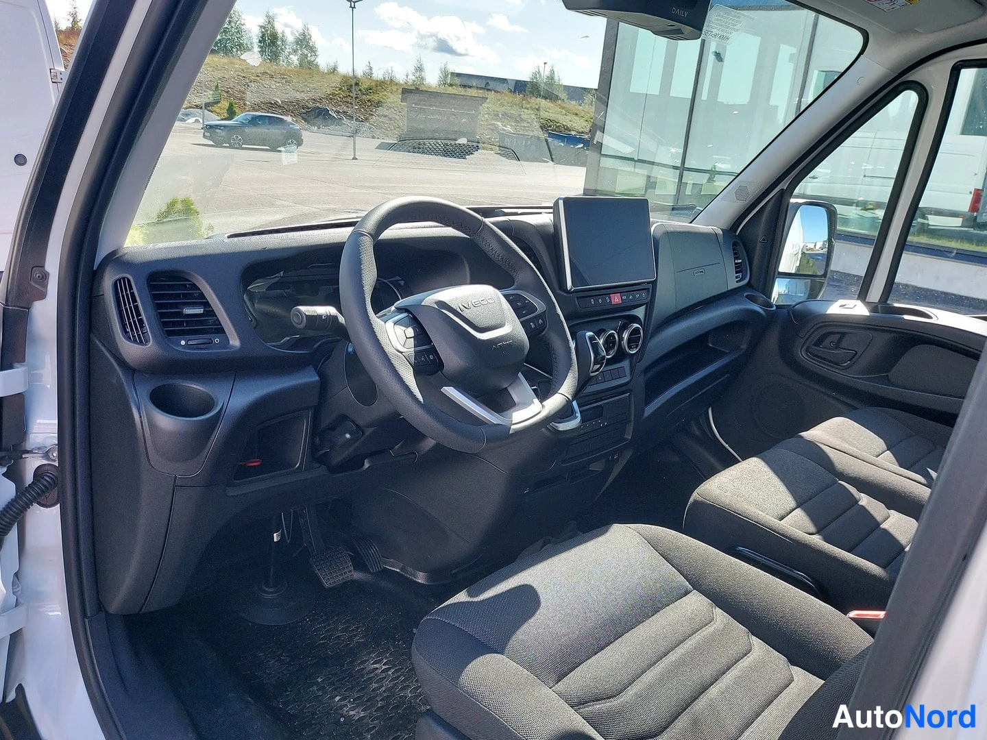Ohjaamo: Cabin view of the Iveco Daily interior showing a two-bench seat layout, with a driver's seat on the left and a passenger bench on the right. The dashboard featu