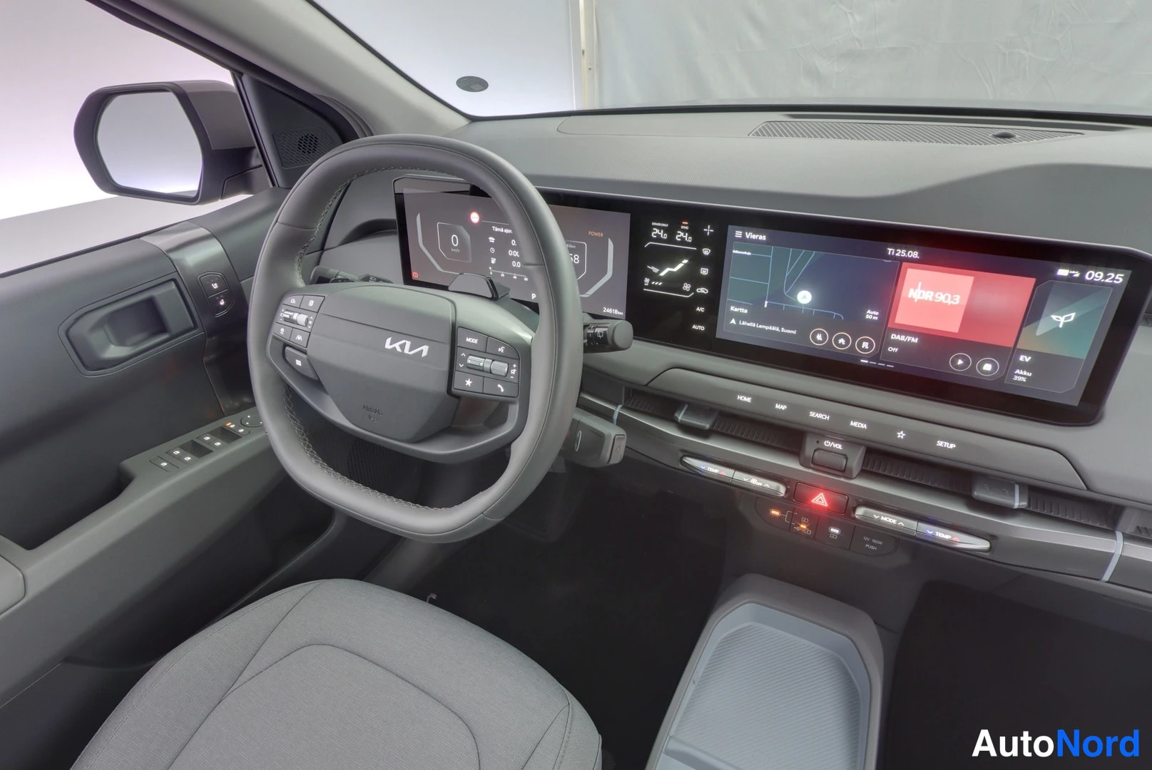 Edestä: Kia EV3:n ohjaamo kuljettajan oven puolelta: KIA-merkkinen ohjauspyörä, laaja näyttöpaneeli ja ovipaneelin säätimet.