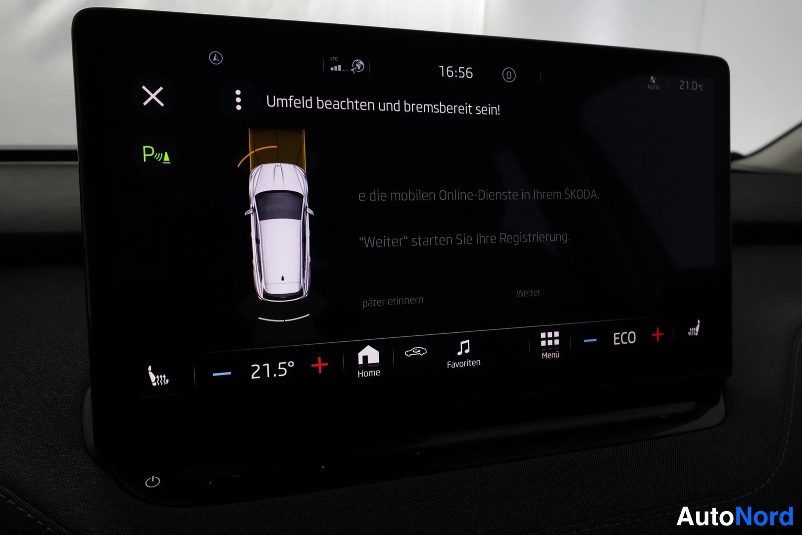 Muu: Kosketusnäytön infotainment-järjestelmä näyttää pysäköintianturinäkymän autosta ylhäältä ja saksankielisen tekstin 'Umfeld beachten und bremsbereit sein!'.