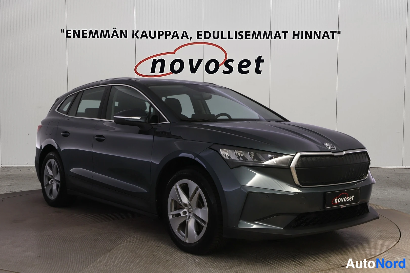 Etuviisto, nokka oikealle: Tummanharmaa Skoda Enyaq kuvattuna etuviistosta, nokka oikealla: etusäleikkö, ajovalot ja vasen kylki näkyvät, taustalla valkoinen seinä novoset-tekstill