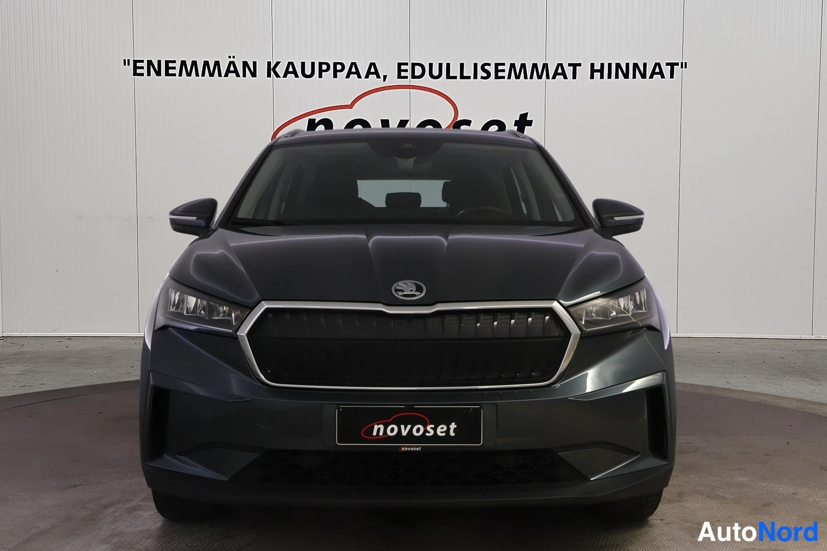 Edestä: Skoda Enyaq suoraan edestä kuvattuna: tumma etusäleikkö, ajovalot ja etupuskuri näkyvät, taustalla valkoinen seinä novoset-tekstillä.