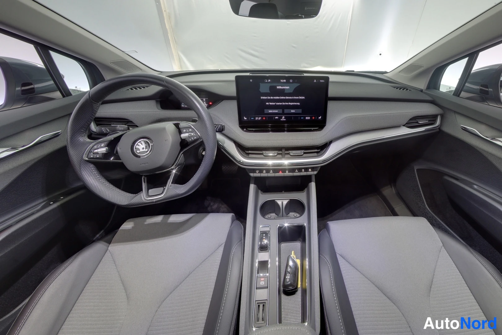 Muu: Auton ohjaamo ylhäältä kuvattuna: Skoda-ohjauspyörä, keskellä infotainment-näyttö ja vaakasuora ilmastointiritilä, kaksi tummaa etuistuinta, keskikonsolissa kup