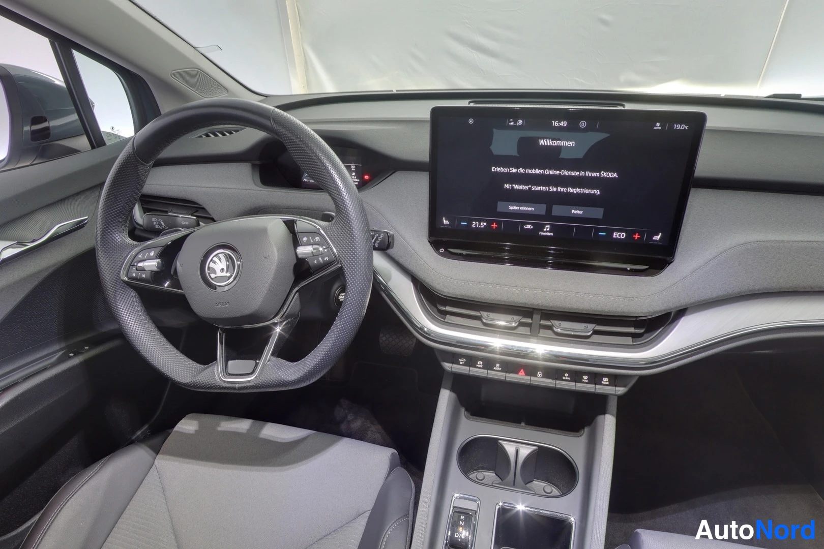Muu: Ohjaamon kojelauta lähikuvassa vasemmalta: Skoda-ohjauspyörä ja infotainment-näyttö, jonka Willkommen-ruudussa saksankielinen teksti ja aika 16:49.