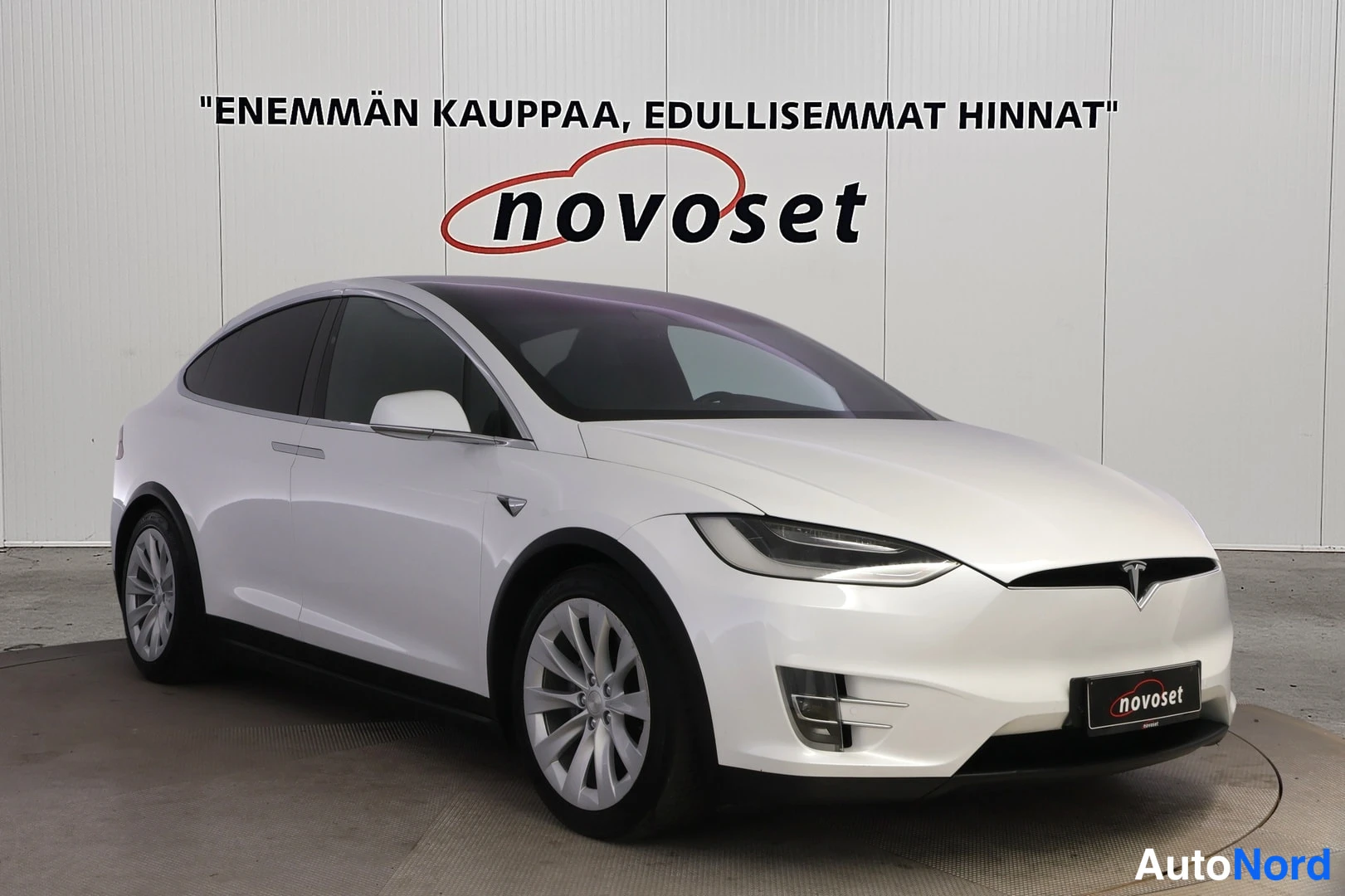 Etuviisto, nokka oikealle: Valkoisen Tesla Model X:n etuosan ja vasemman sivun kolmasosan näkymä studiossa, Novoset-kyltti taustalla.
