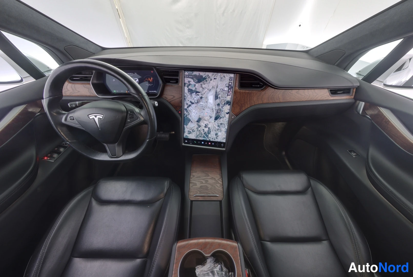 Muu: Mustapenkkisen Tesla Model X:n ohjaamo ylhäältä: puukoristeinen kojelauta, ohjauspyörä ja suuri keskikosketusnäyttö karttaan.