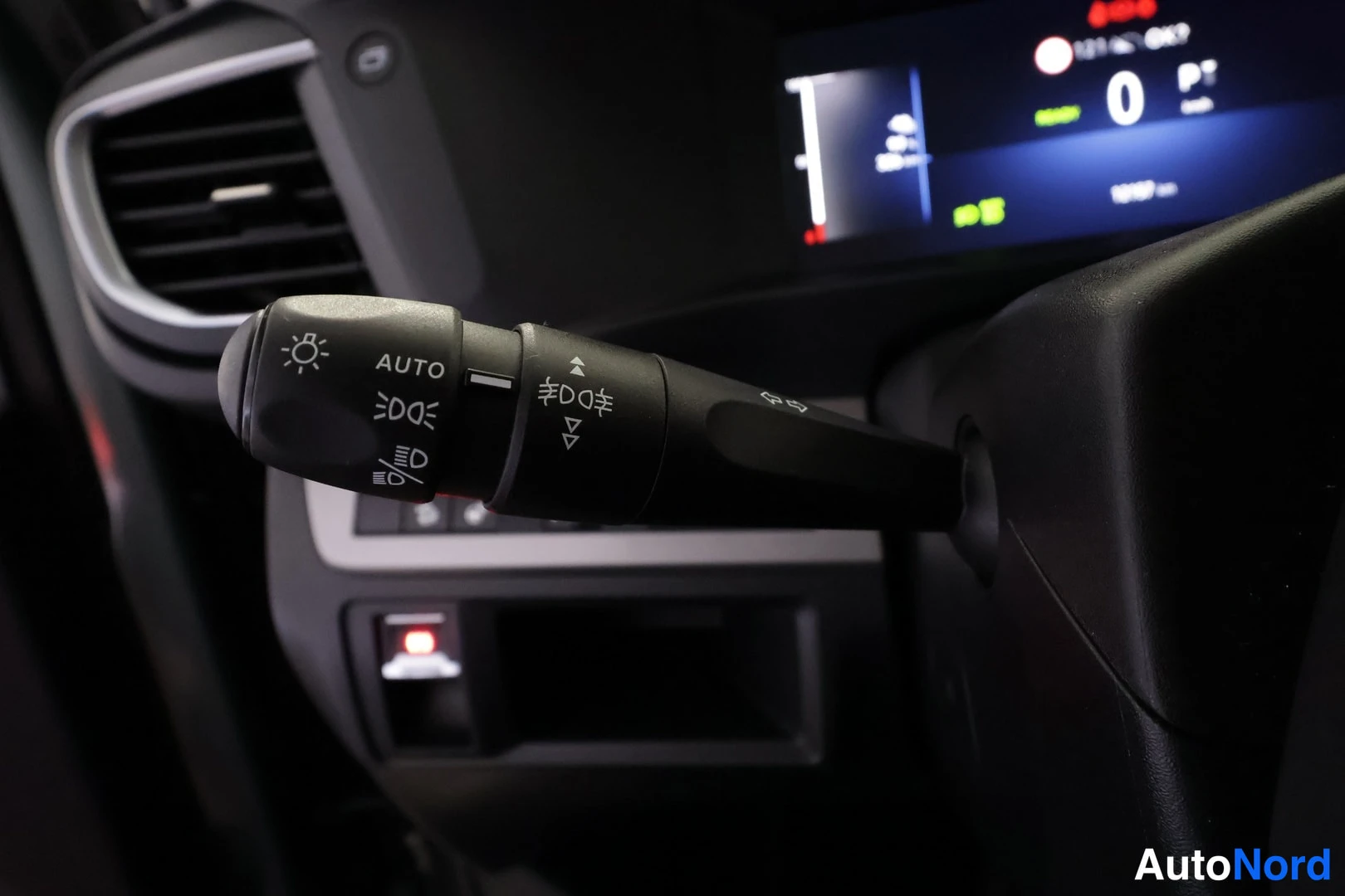 Yksityiskohta: Valokytkimen vivut AUTO-merkinnällä, taustalla kojelaudan näyttö ja ilmastointilaitteet.