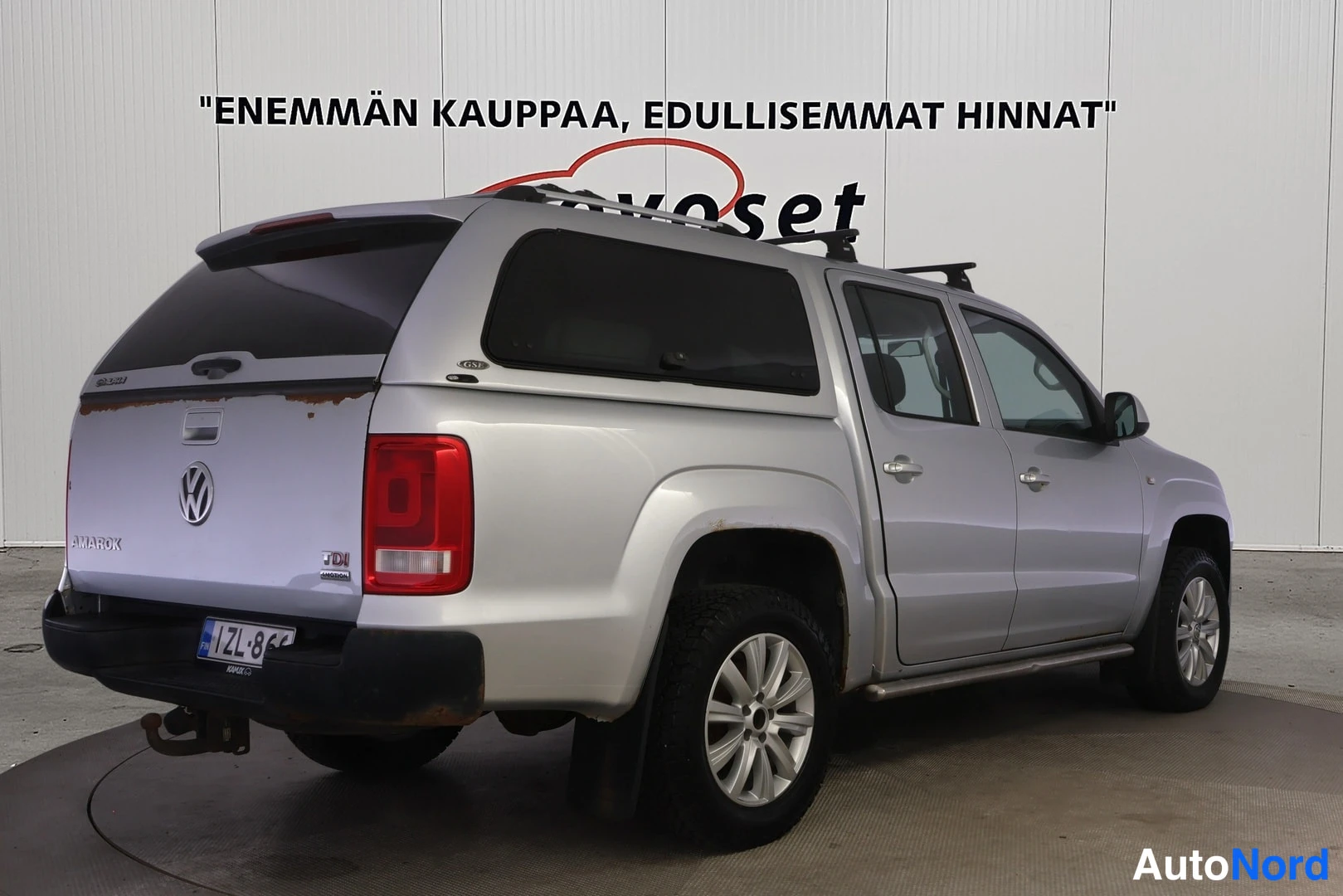 Takaviisto: Hopeanvärinen Volkswagen Amarok takaviistosta vasemmalta, kuorma-A-kansi, vetokoukku ja jalkalaudat näkyvissä.